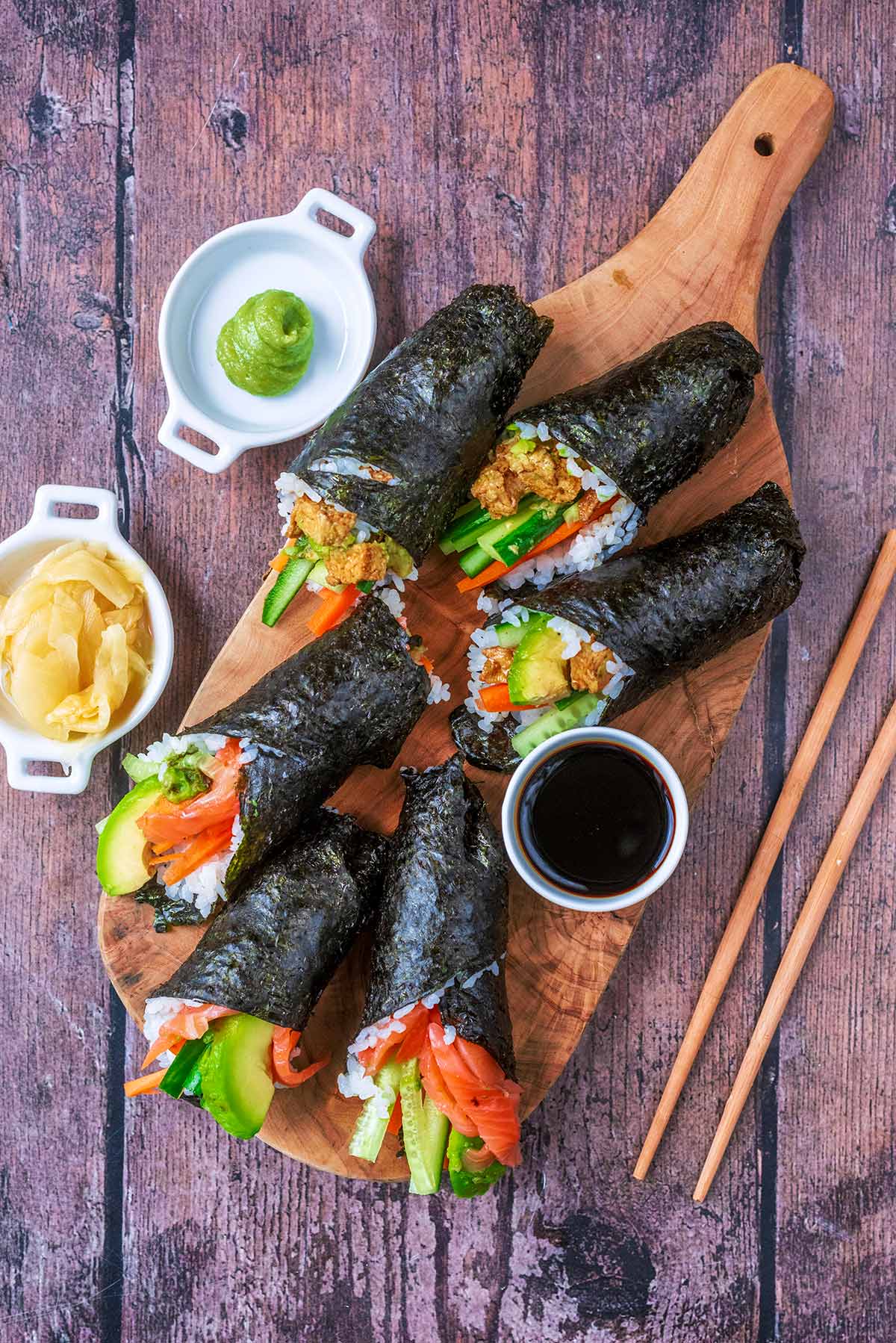 Six sushi hand rolls on a wooden board with pots of soy sauce, ginger and wasabi.