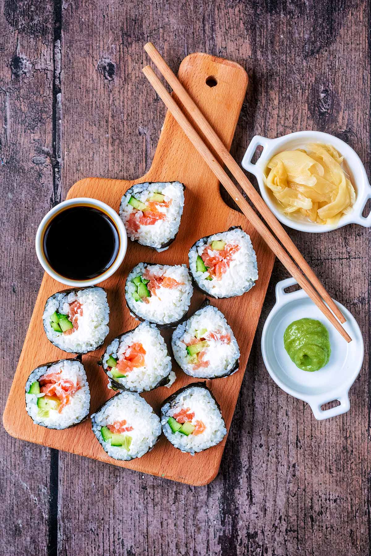 Pieces of cream cheese sushi on a wooden board with pots of soy sauce, wasabi and ginger.