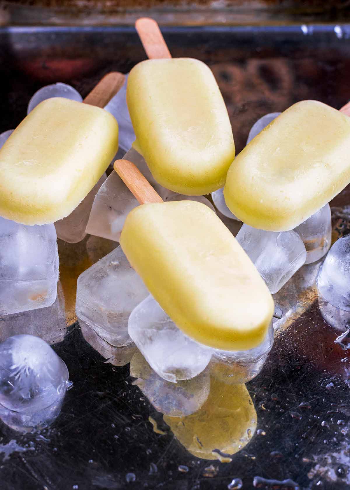 Four pineapple ice lollies on a tray surrounded by ice cubes.