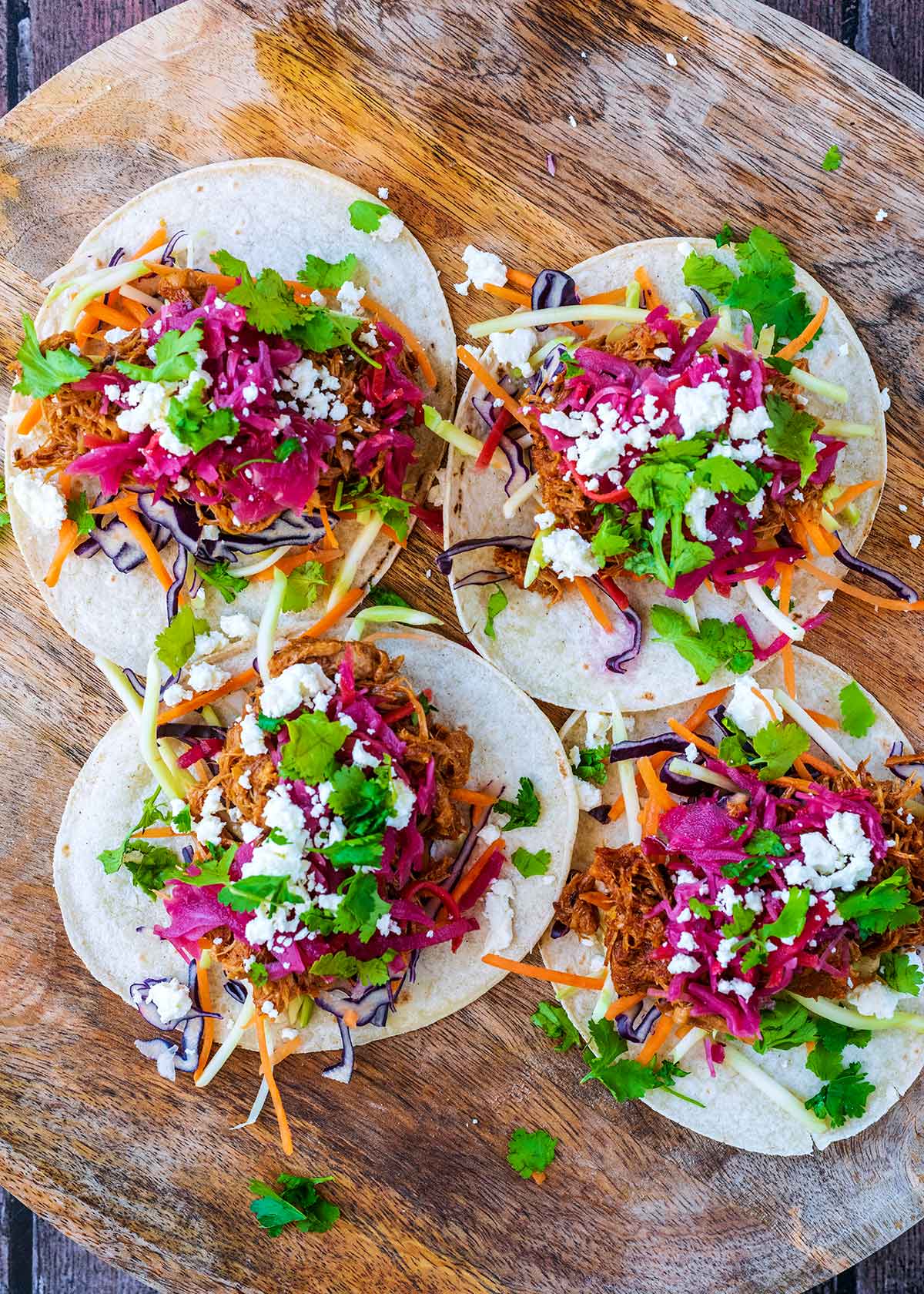 Four open pork and vegetable tacos on a round wooden board.