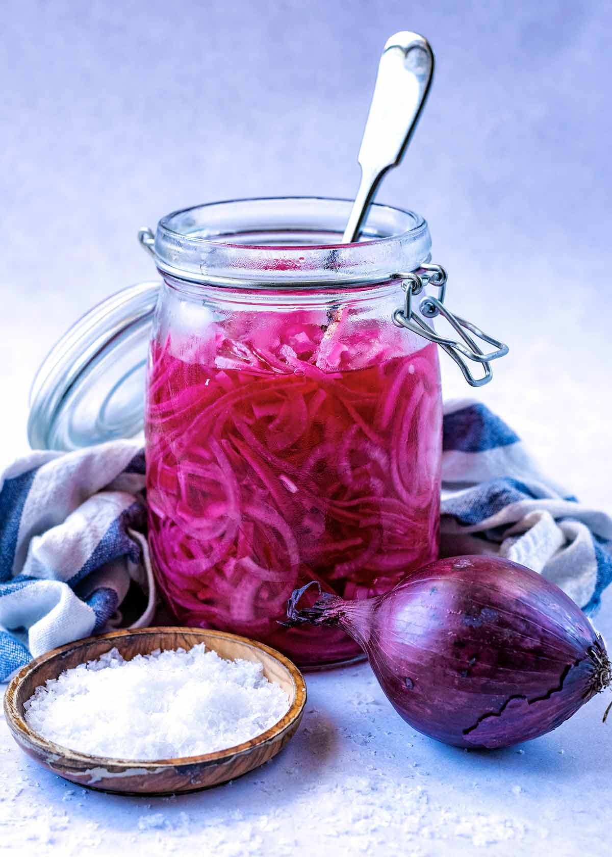 A jar of sliced pickled onions next to an onion and a small dish of salt.
