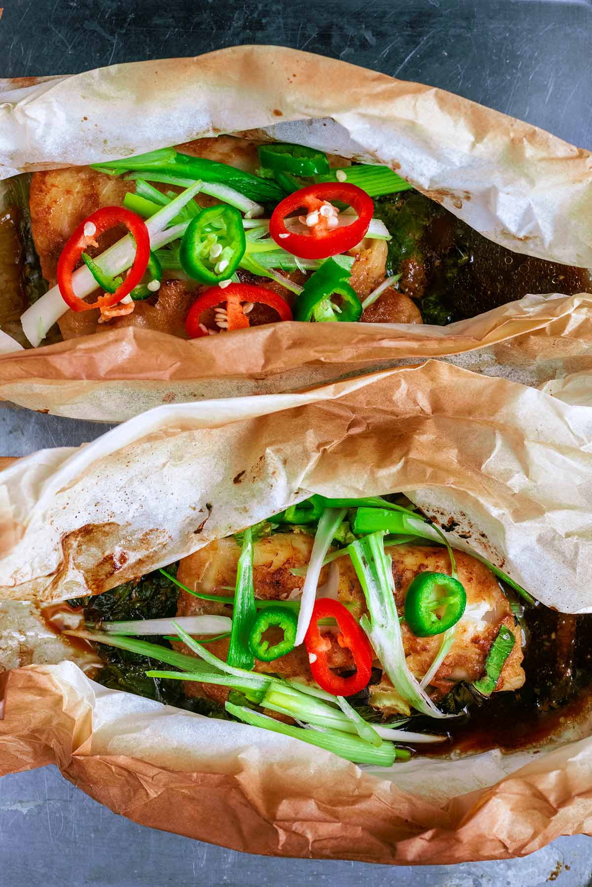 Two open parchment paper parcels containing fish, onions, chillies and a dark sauce.