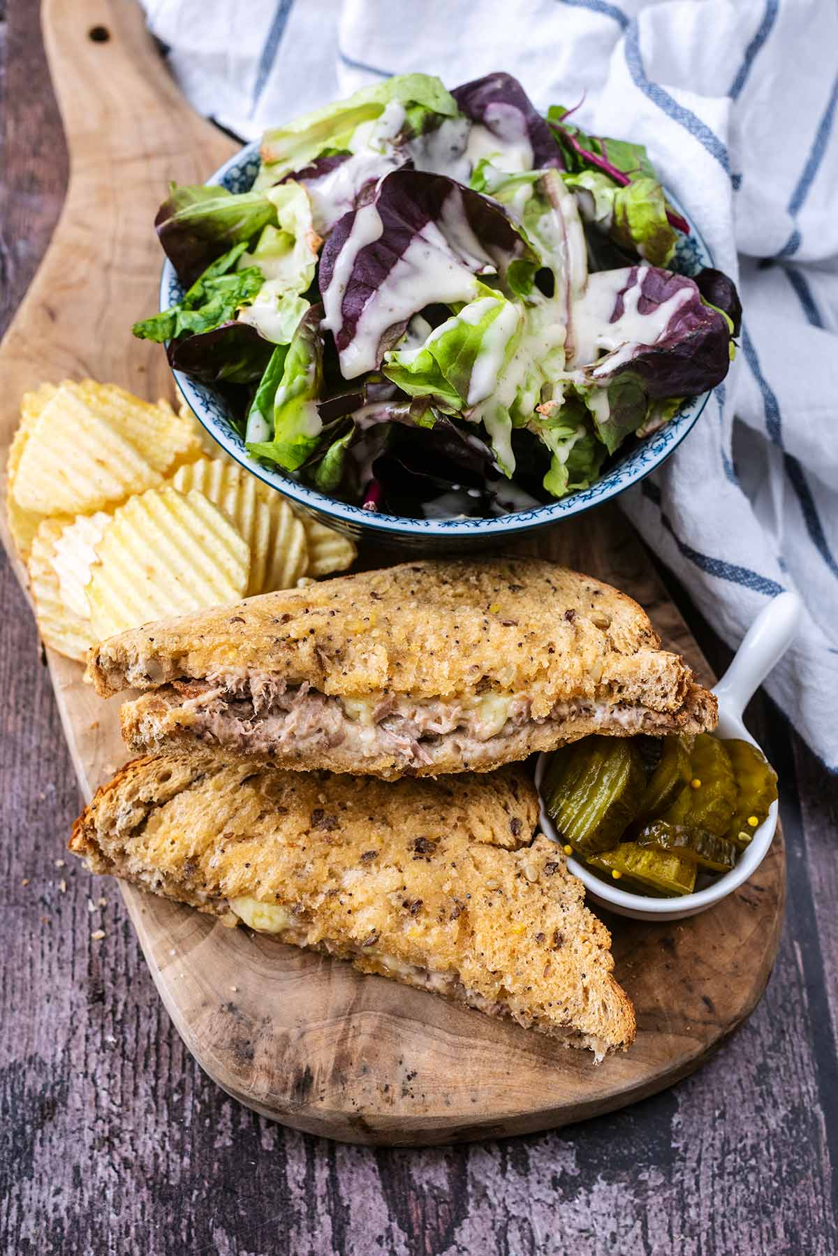 Toasted tuna and cheese sandwich on a wooden board with a bowl of salad.