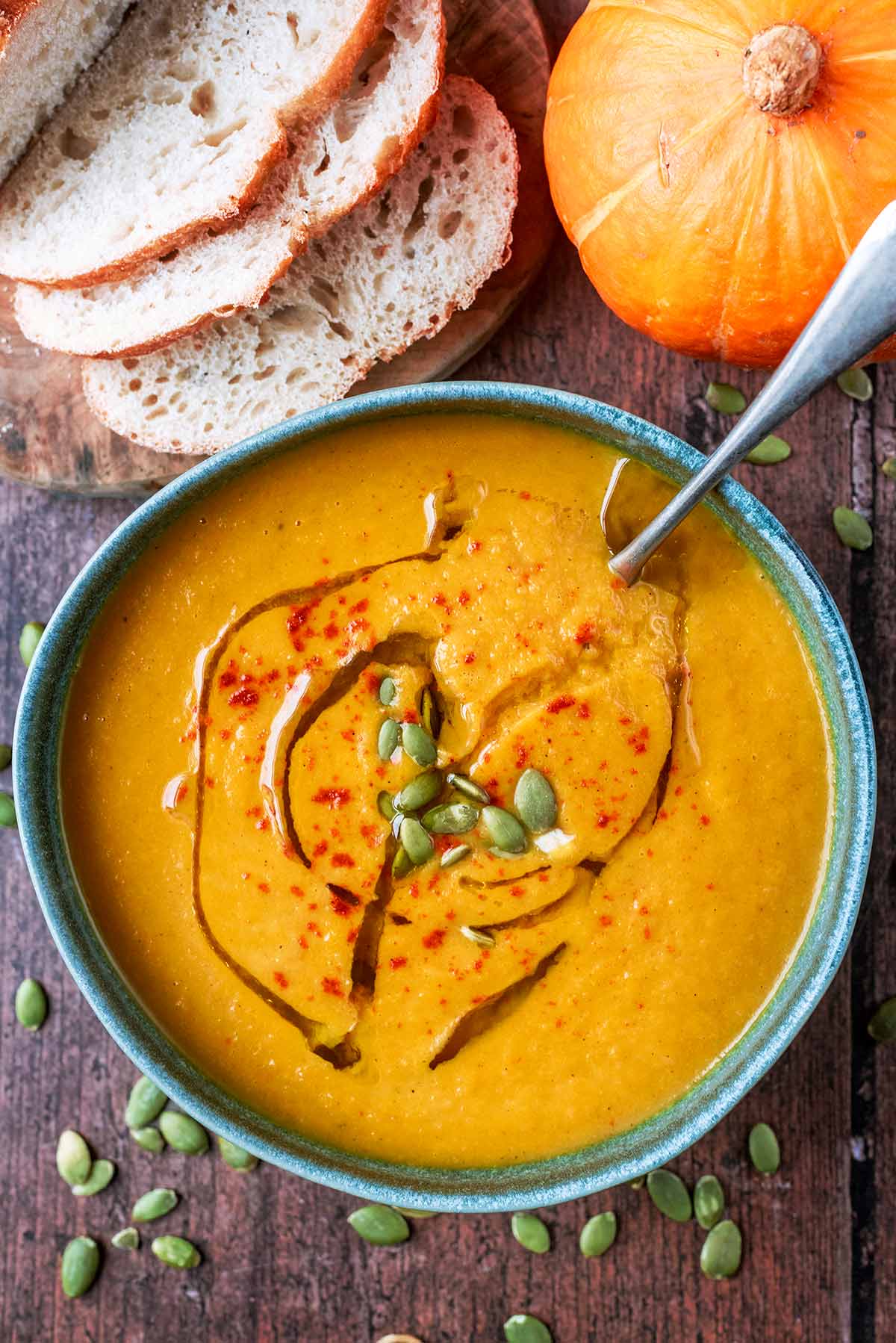 A bowl of pumpkin soup next to a whole pumpkin and a loaf of bread.