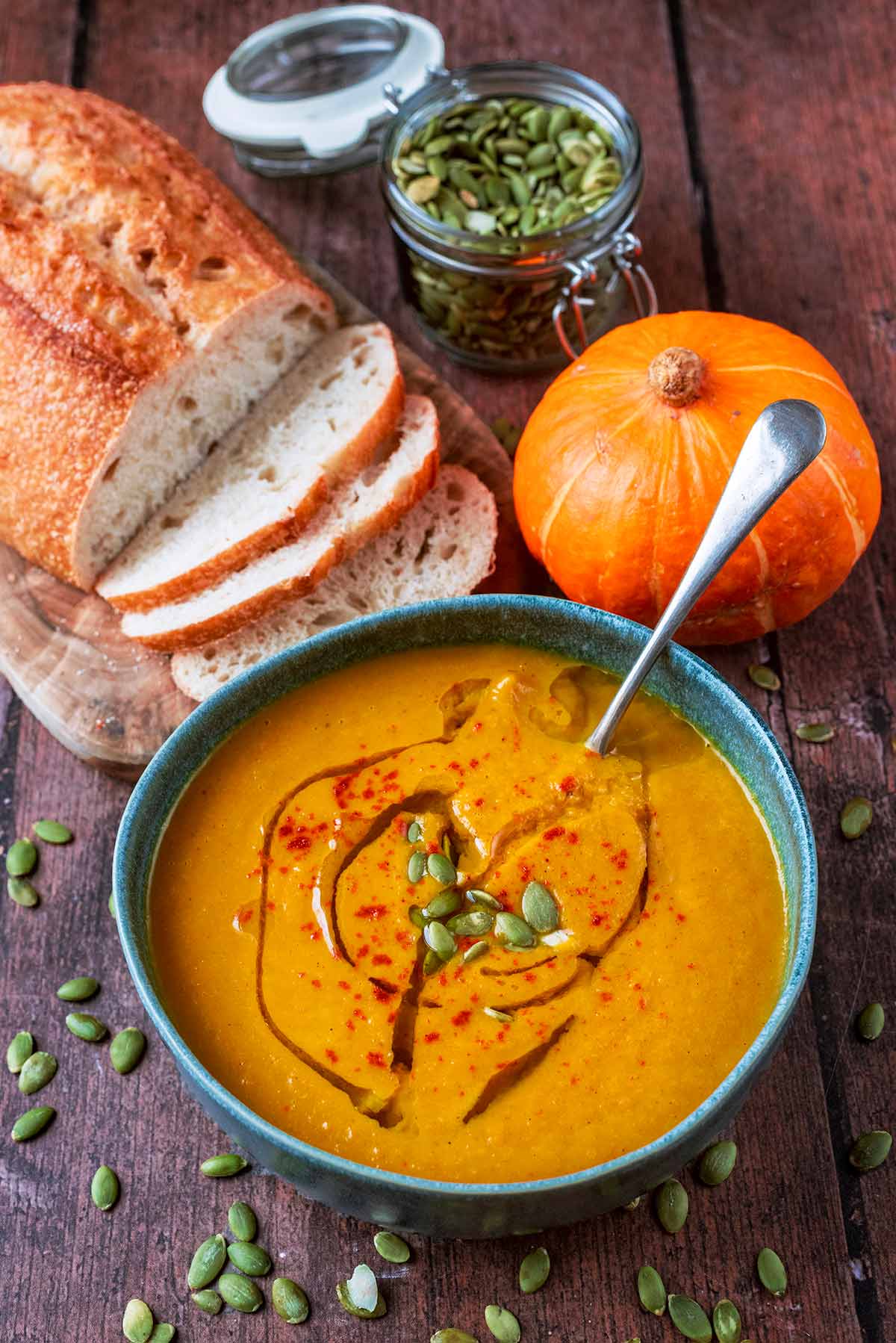 A bowl of pumpkin soup in front of a pumkin, a jar of pumpkin seeds and some bread.