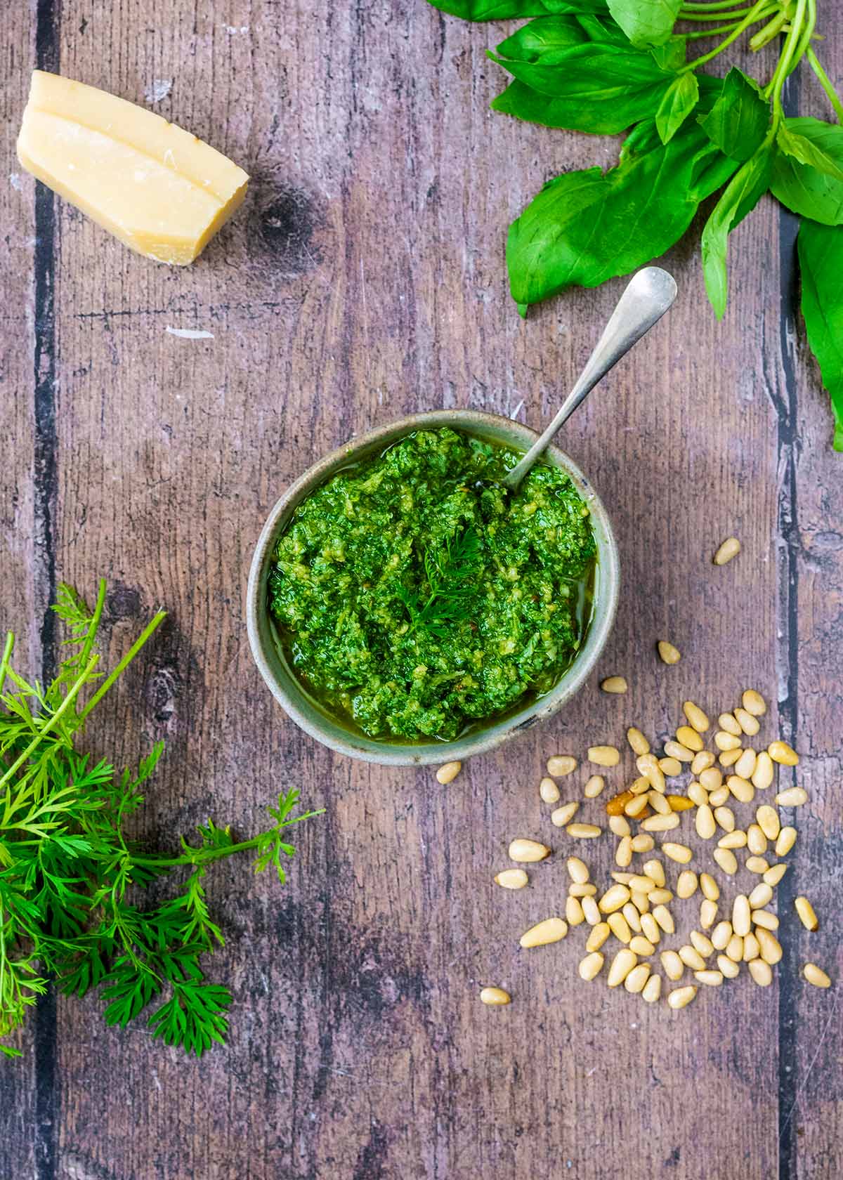 A bowl of pesto surrounded by parmesan cheese, basil leaves, pine nuts and carrot tops.