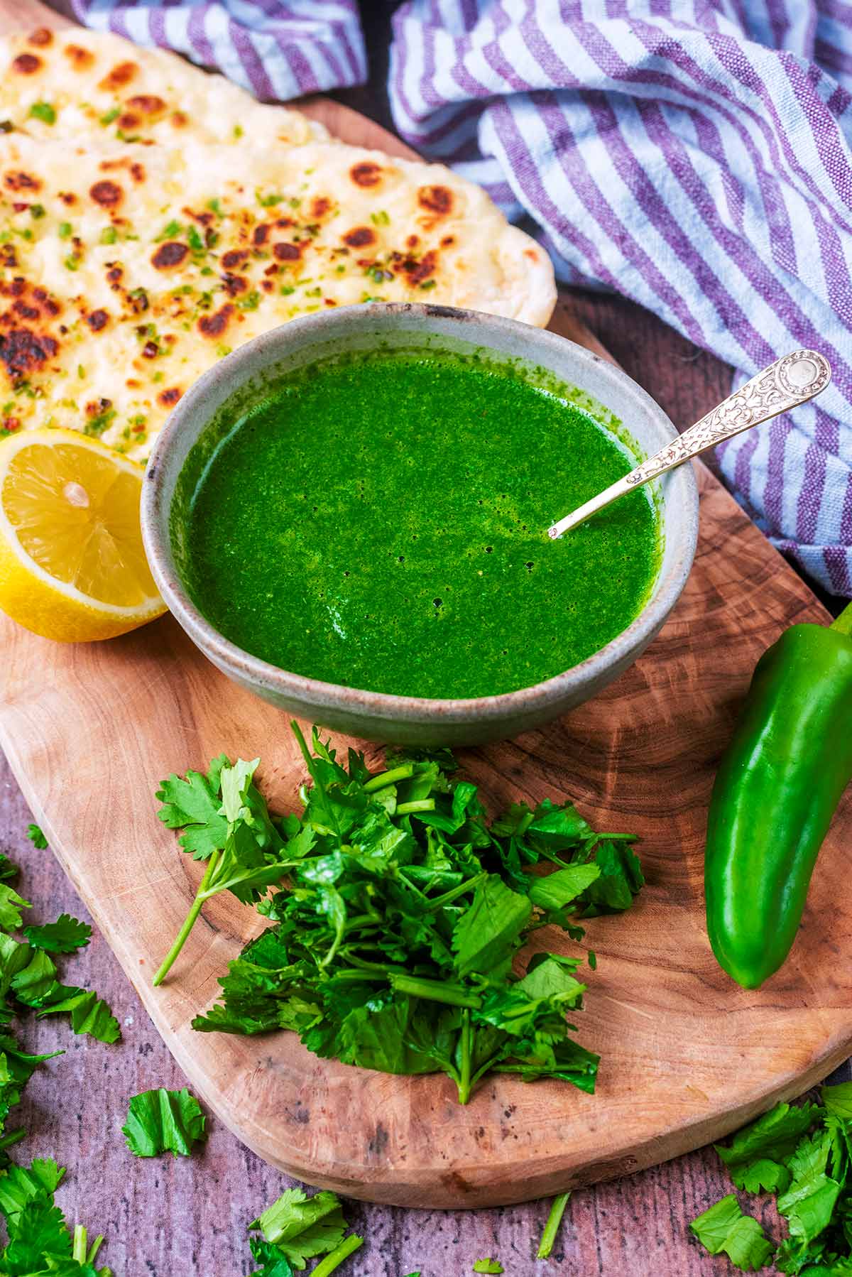 A bowl of green sauce on a board with coriander leaves and a naan bread.