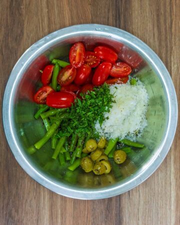 A bowl containing cooked green beans, tomatoes, olives, grated cheese and chopped mint.