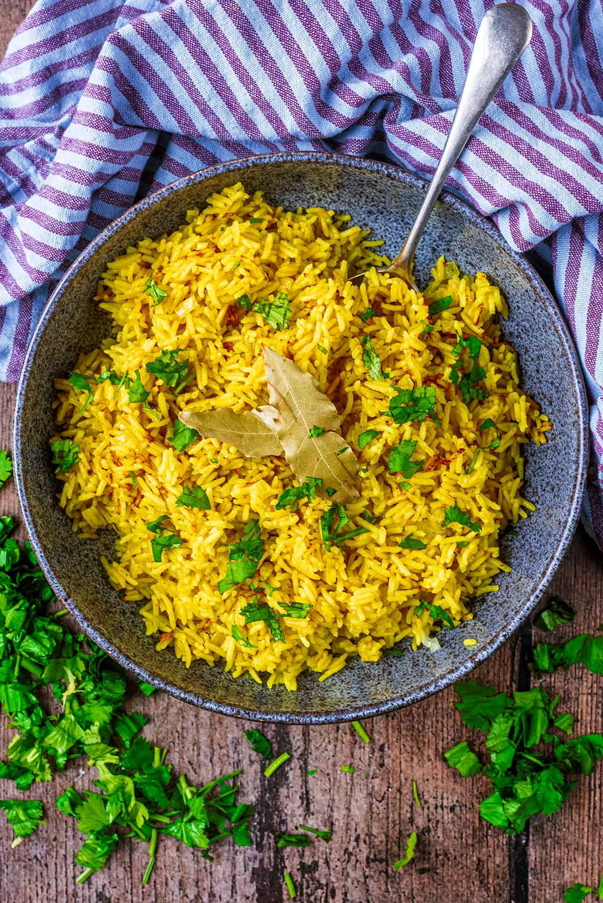 A bowl of yellow coloured rice next to a striped towel and chopped coriander.