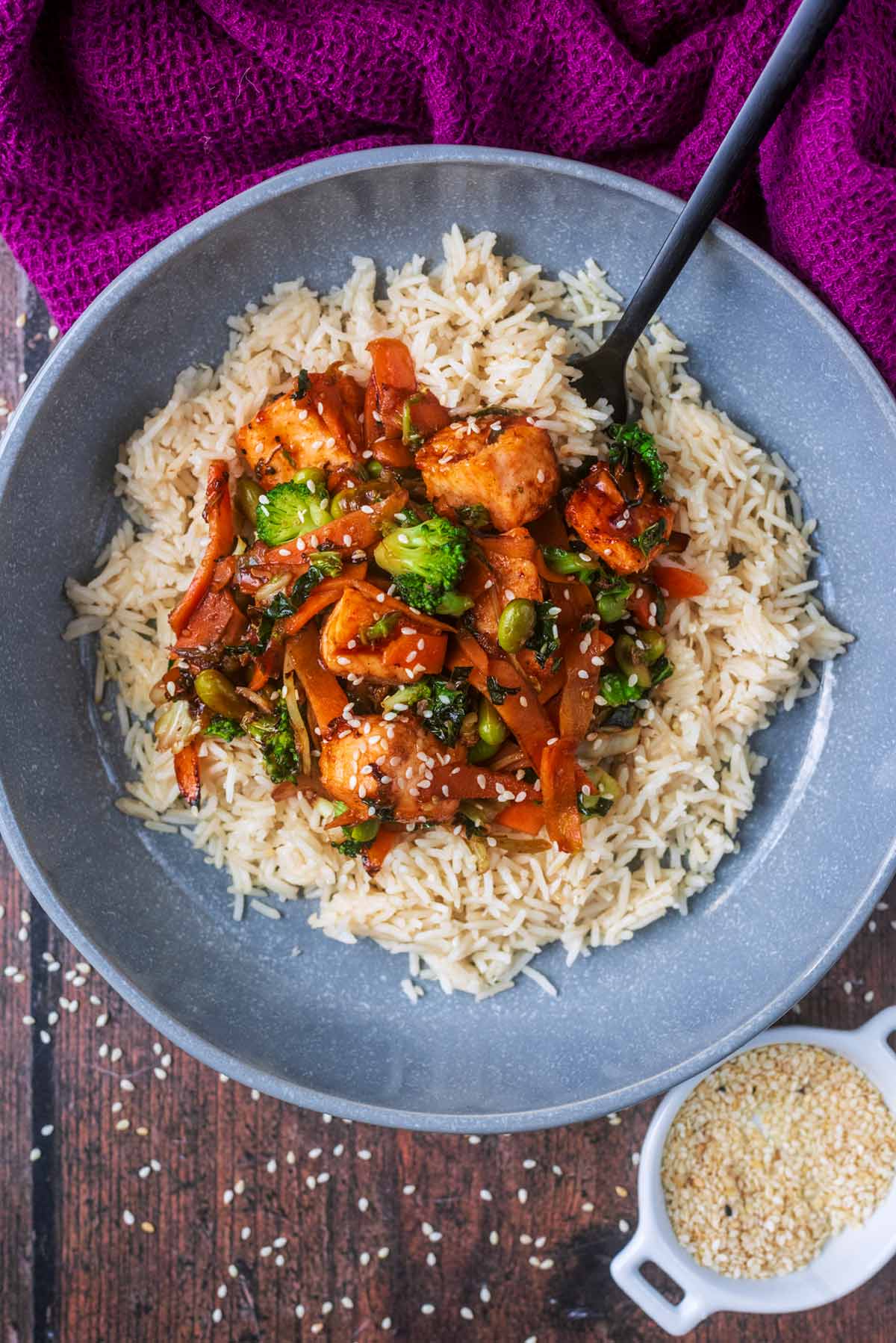Salmon stir fry surrounded by cooked rice in a grey bowl.