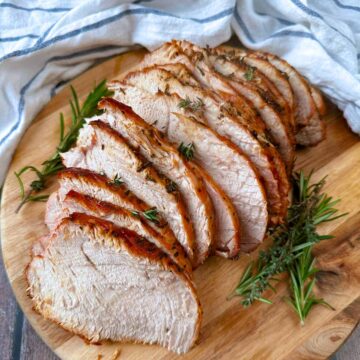 Slices of air fryer turkey on a wooden board with sprigs of rosemary and thyme.