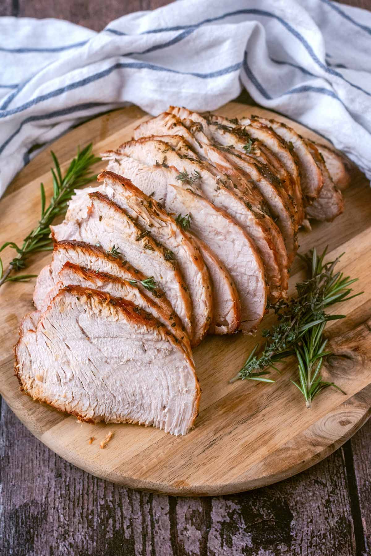 Sliced cooked turkey on a wooden serving board in front of a striped towel.