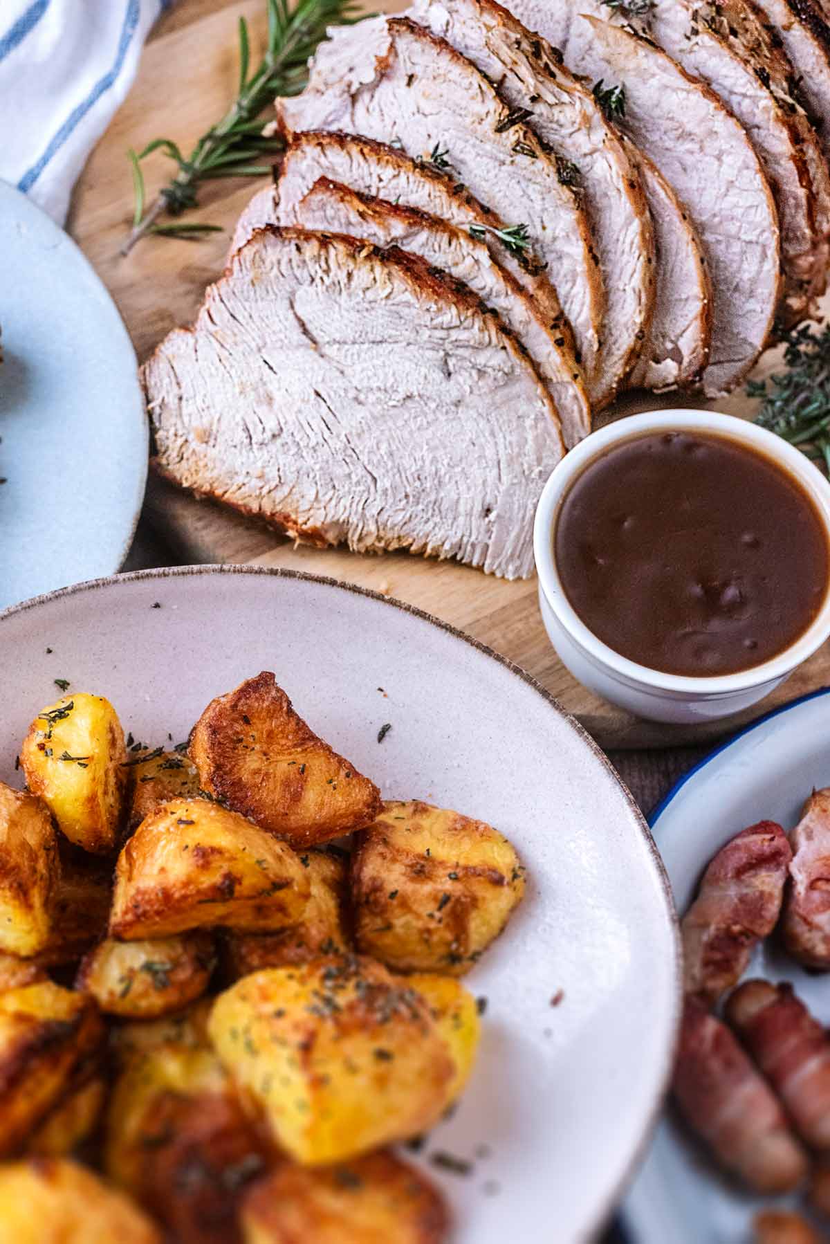 Slices of cooked turkey next to some roast potatoes, pigs in blankets and some gravy.