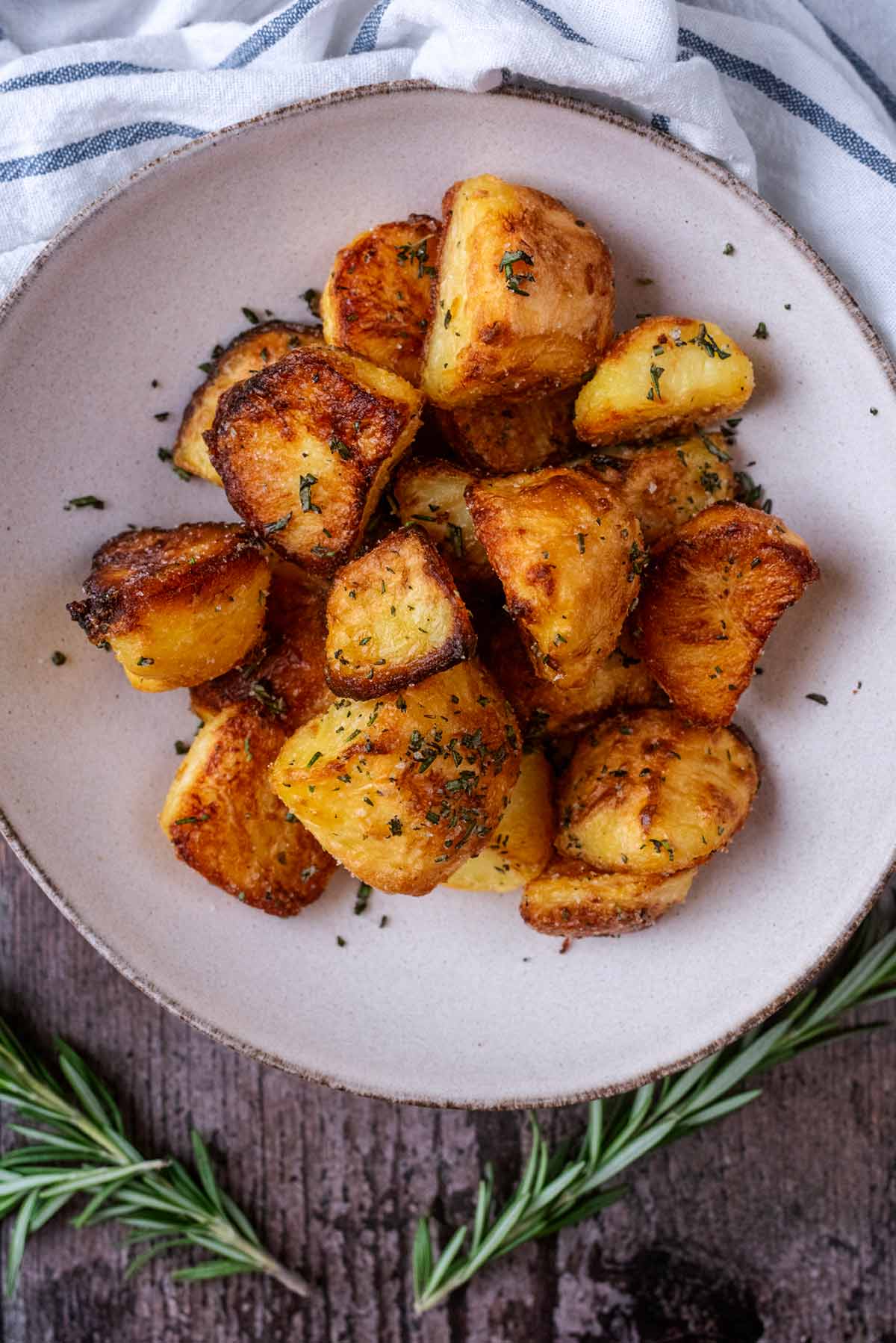 A bowl of roast potatoes next to a striped towel and some sprigs of rosemary.