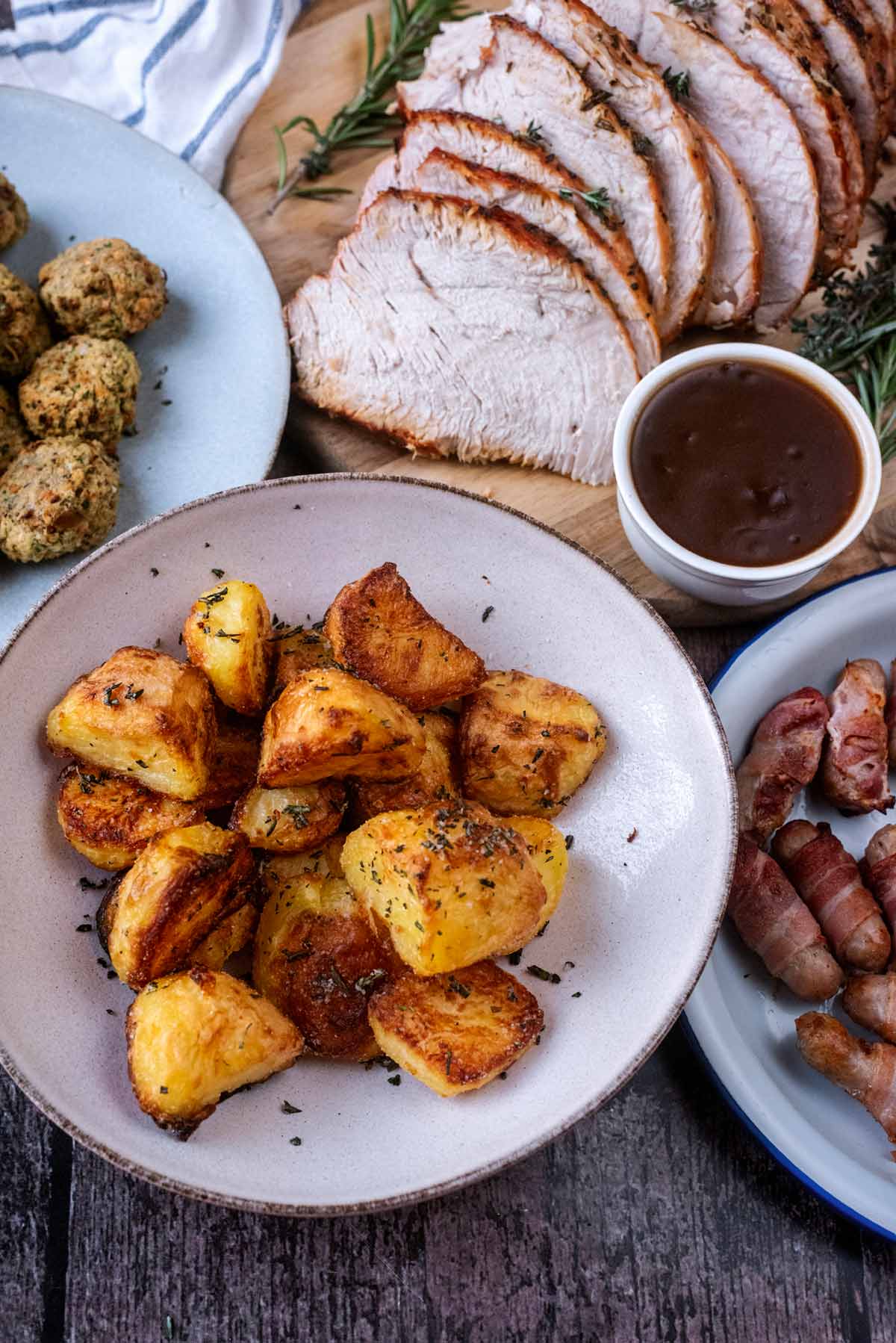 A bowl of roast potatoes in front of some sliced turkey and a small pot of gravy.