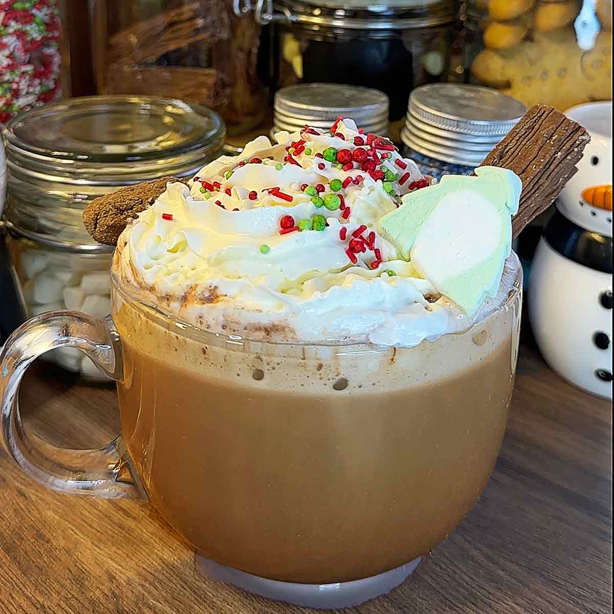 A glass mug full of hot chocolate in front of a hot chocolate station.