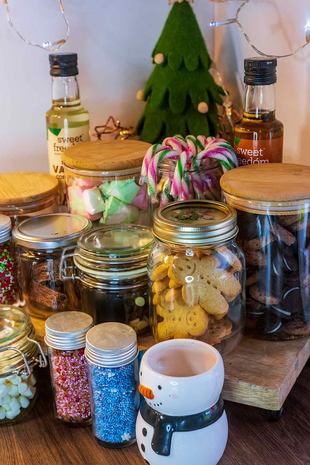 A snowman shaped mug in front of a jar of gingerbread men and jars of sugar sprinkles.