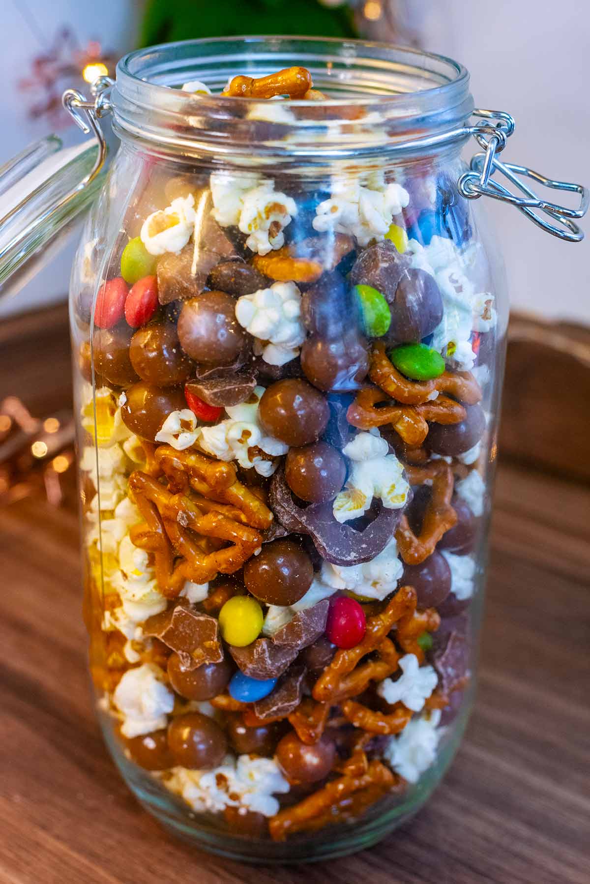 A tall glass jar full of popcorn, pretzels, smarties and maltesers.