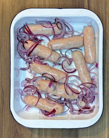 Raw sausages and sliced red onion in a baking dish.