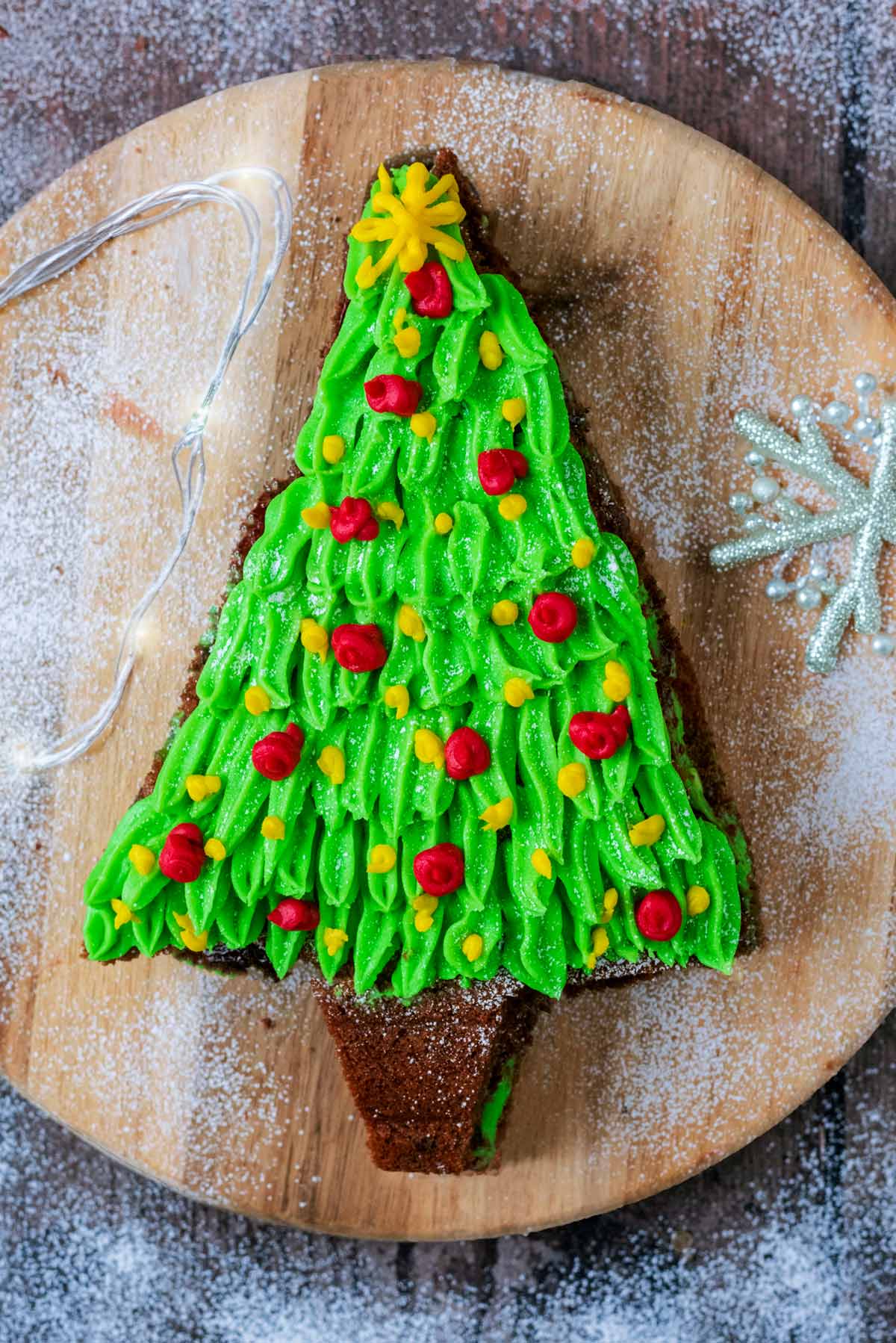 A Christmas tree shaped cake on a board with fairy lights and a decorative snowflake.