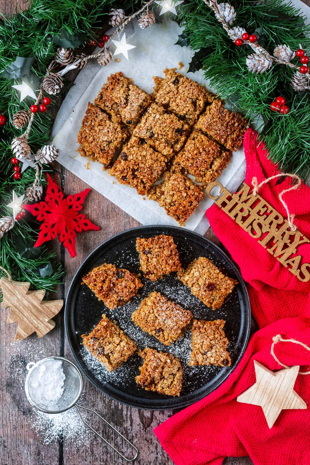 A plate of flapjacks next to flapjacks on some baking paper all surrounded by Christmas decorations.