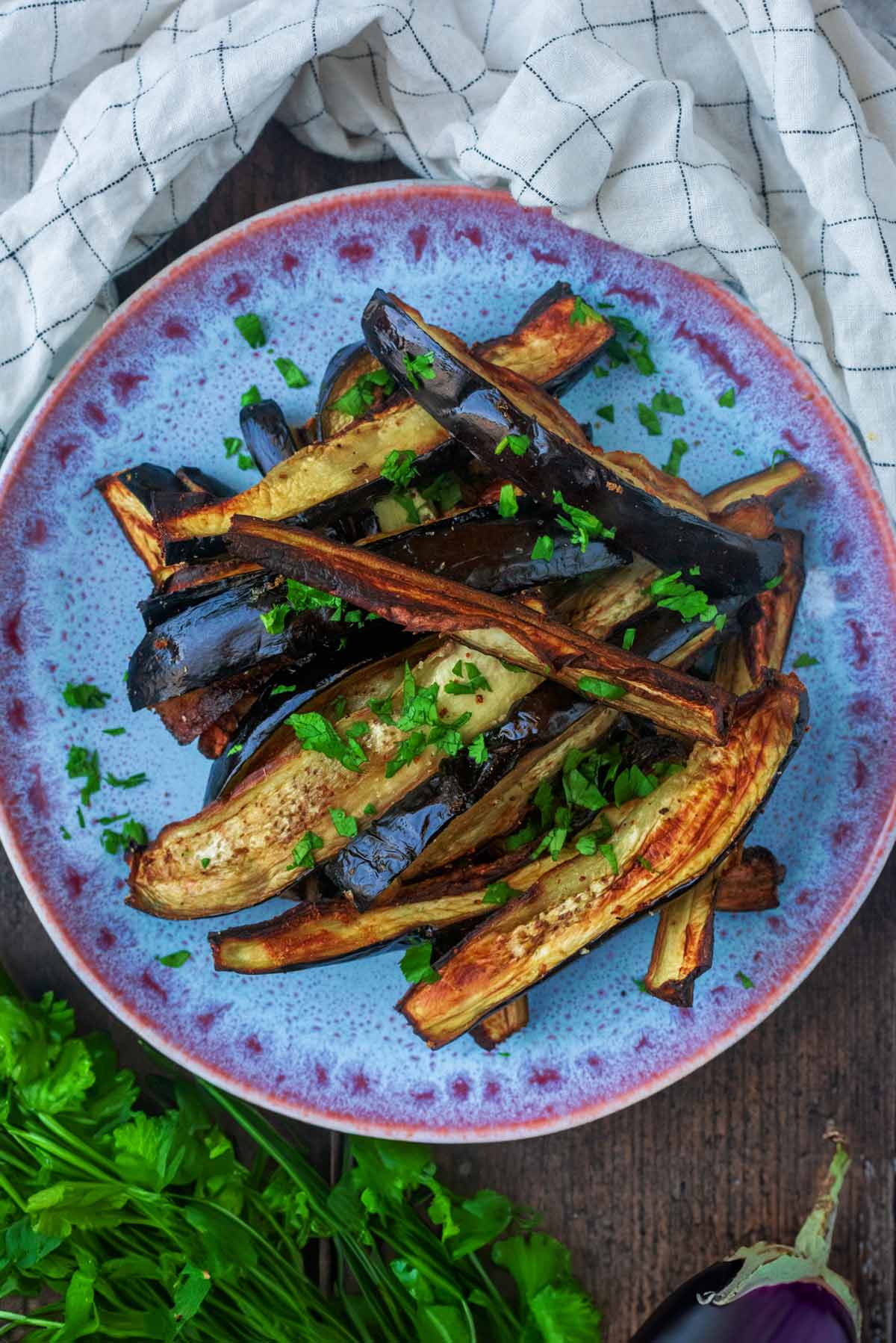 A plate of cooked aubergine strips next to a whole aubergine and a bunch of coriander.