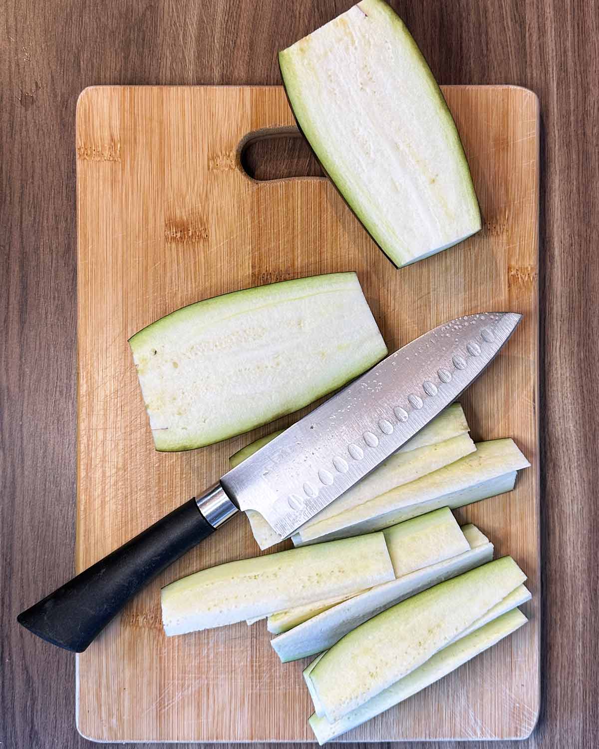 Aubergines being sliced into strips on a wooden chopping board.