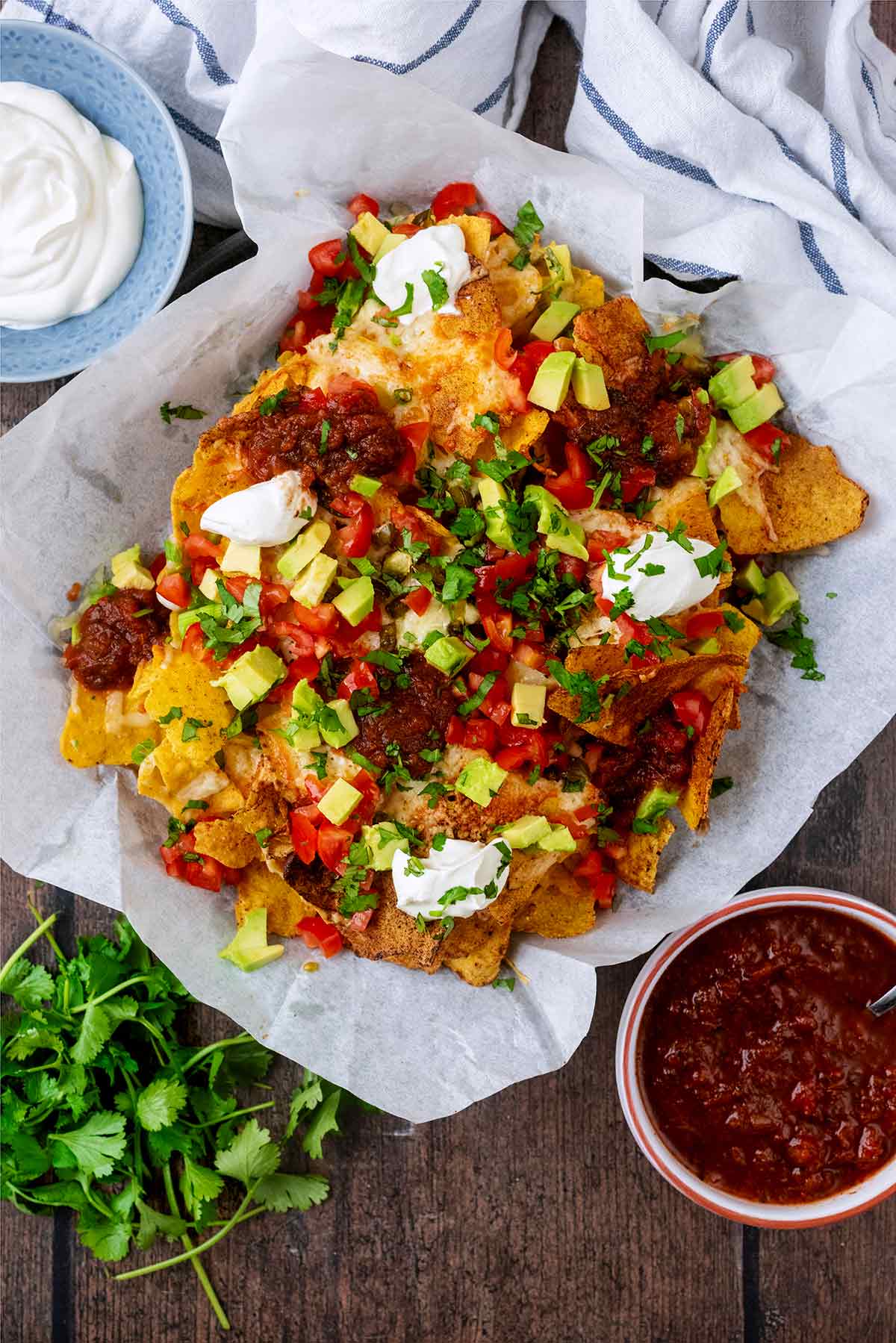 Nachos on a sheet of parchment paper next to a bunch of coriander and a bowl of salsa.