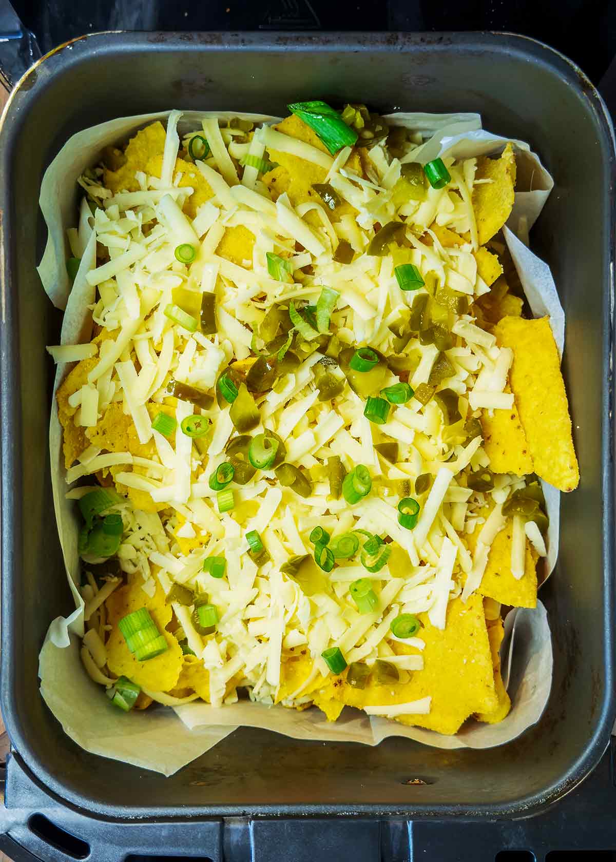 Tortilla chips, cheese, jalapenos and green onions in an air fryer basket.