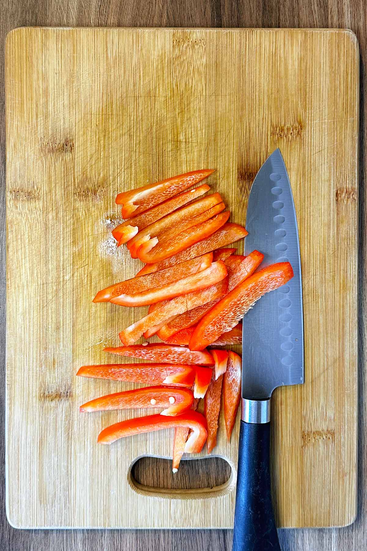 A red bell pepper sliced into strips on a chopping board with a chef's knife.