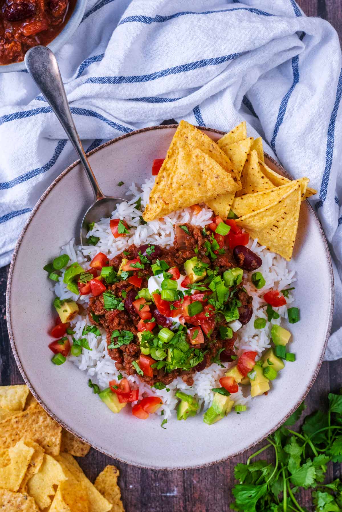 A bowl of chilli and rice next to some tortilla chips and coriander leaves.