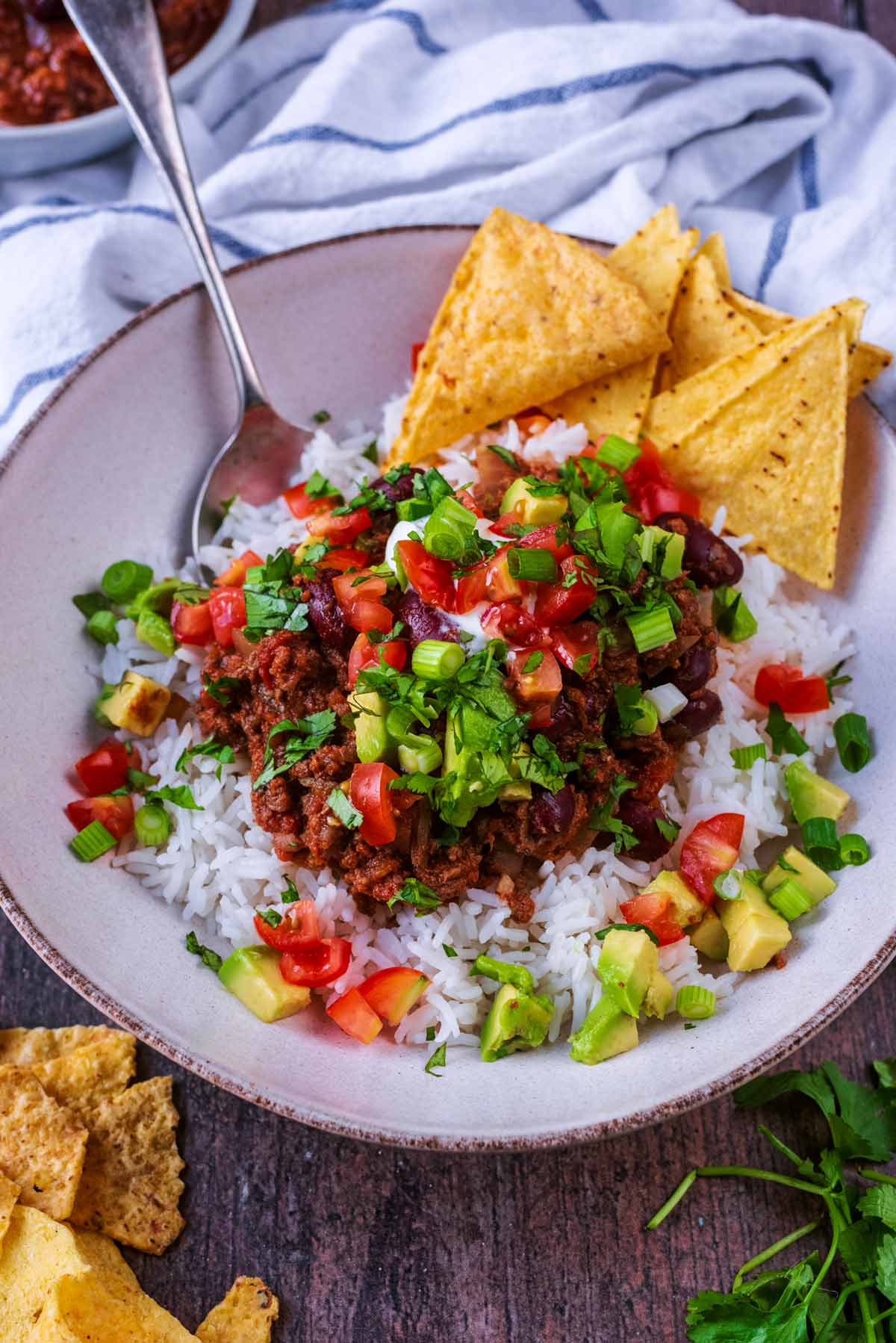 A bowl of chilli and rice with some tortilla chips in front of a striped towel.