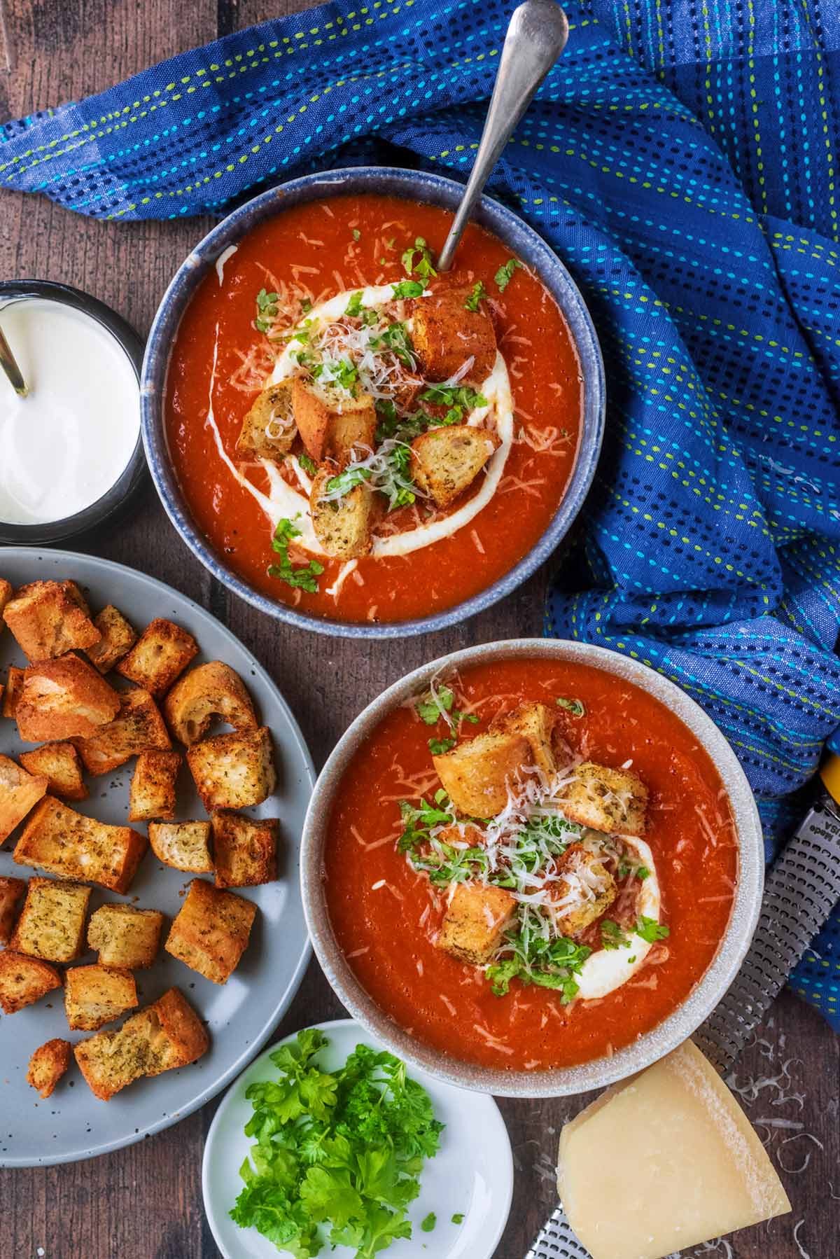 Two bowls of tomato soup next to a plate of croutons, some herbs, cheese and cream.