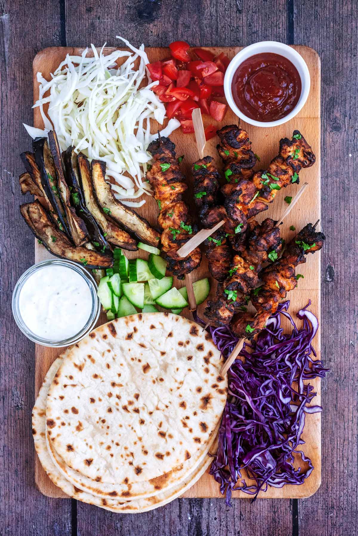 A large serving board with chicken kebabs, flatbreads, vegetables and sauces.
