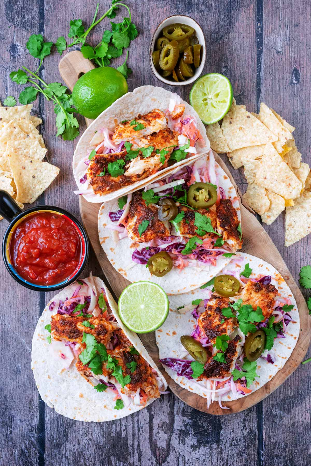 Four fish tacos on a wooden serving board next to salsa, tortilla chips and coriander.