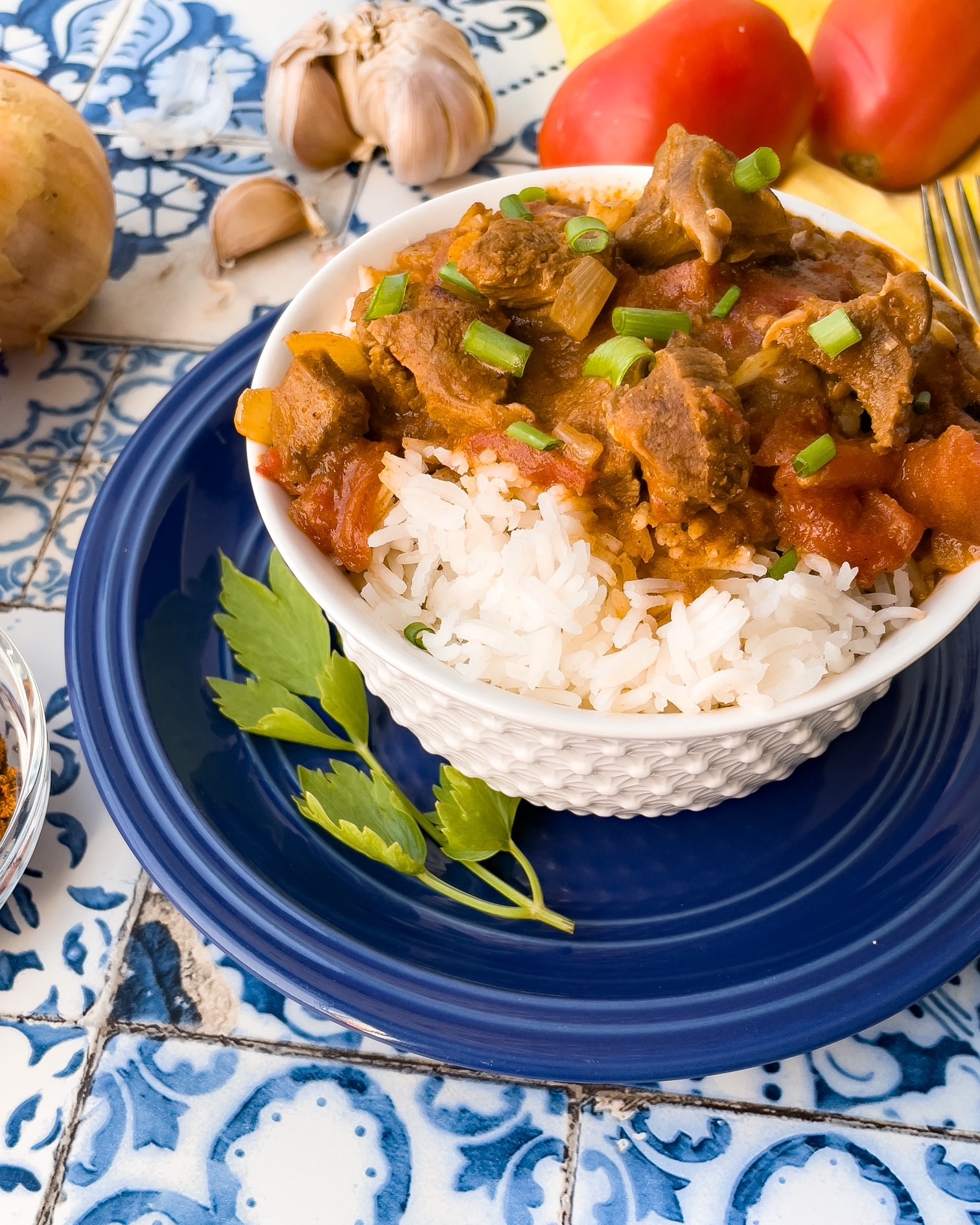 A bowl of lamb curry and rice in front of some tomatoes and a bulb of garlic.
