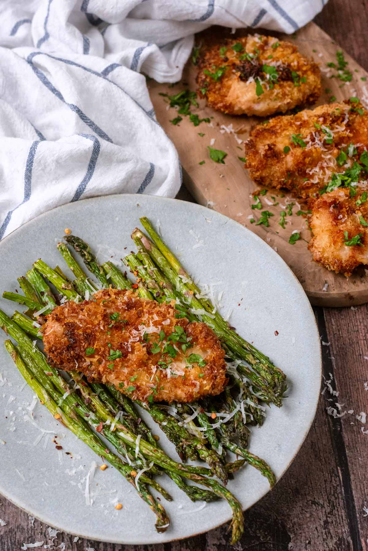 Crusted chicken breast on a plate with some cooked asparagus. More chicken is in the background.