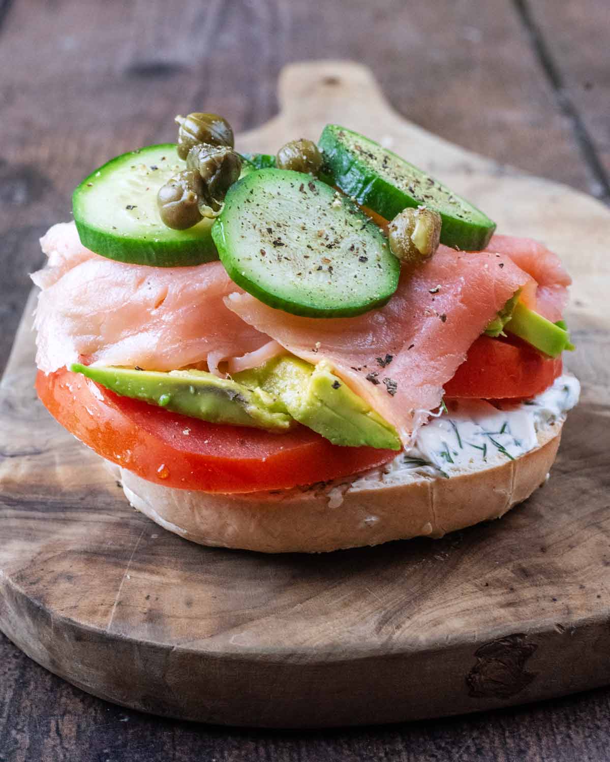 A bagel half topped with cream cheese, tomato slices, salmon, avocado, cucumber slices and capers.