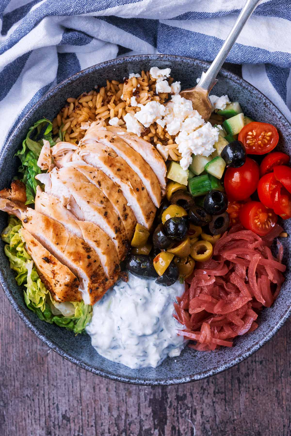 A sliced chicken breast in a bowl with lettuce, rice, tzatziki, tomatoes, olives and feta.