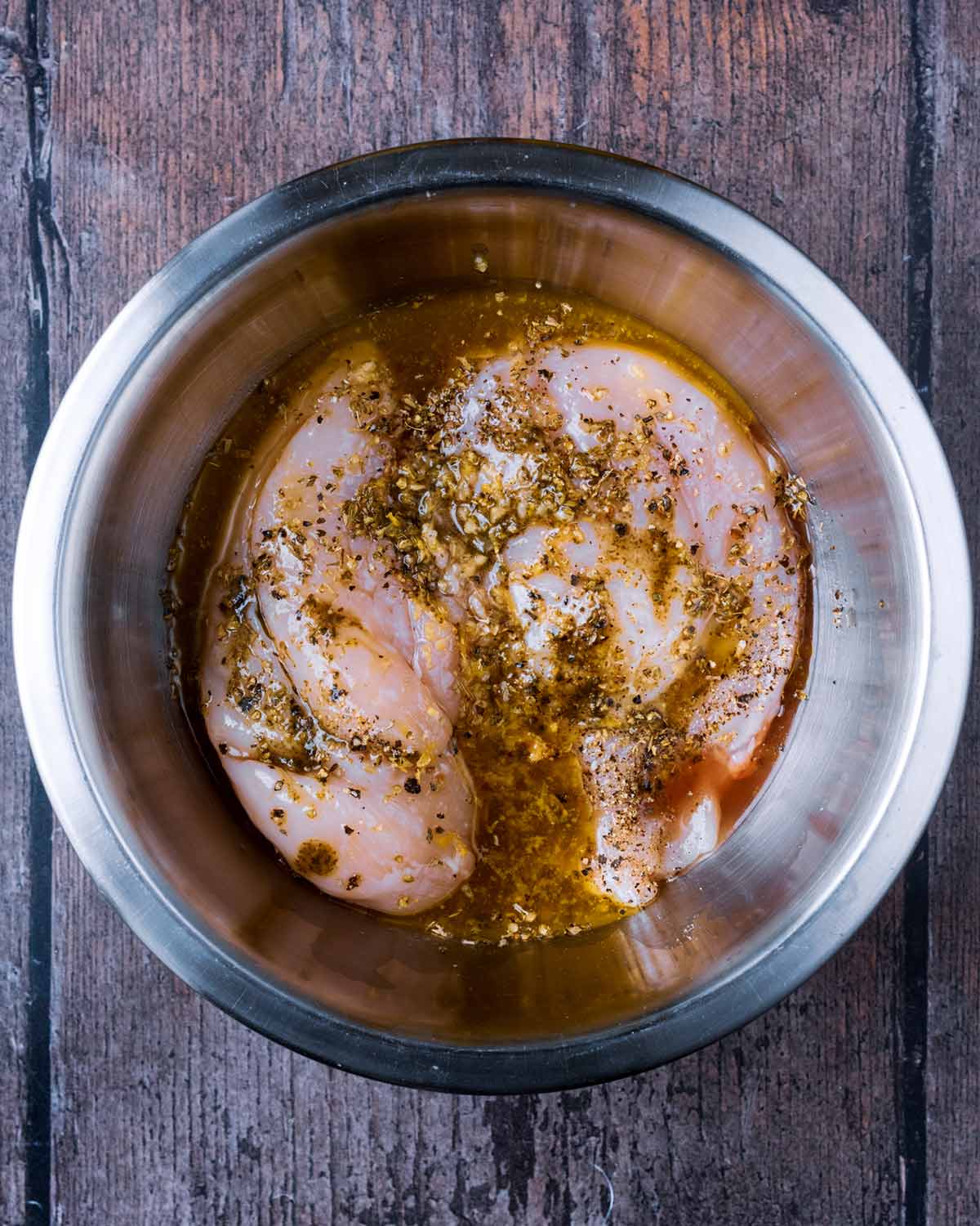 Chicken breasts marinating in oil and herbs in a bowl.