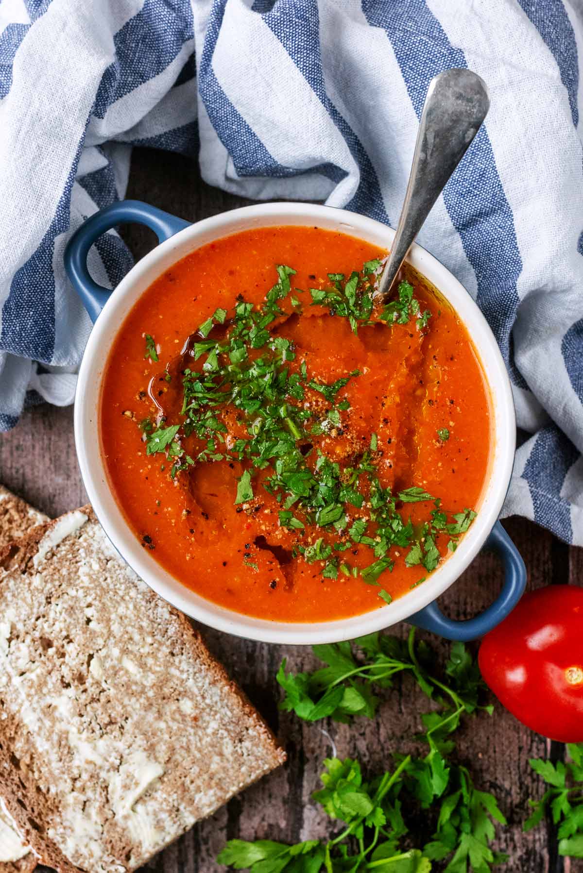 A bowl of red coloured soup next to some buttered bread, herbs and a tomato.