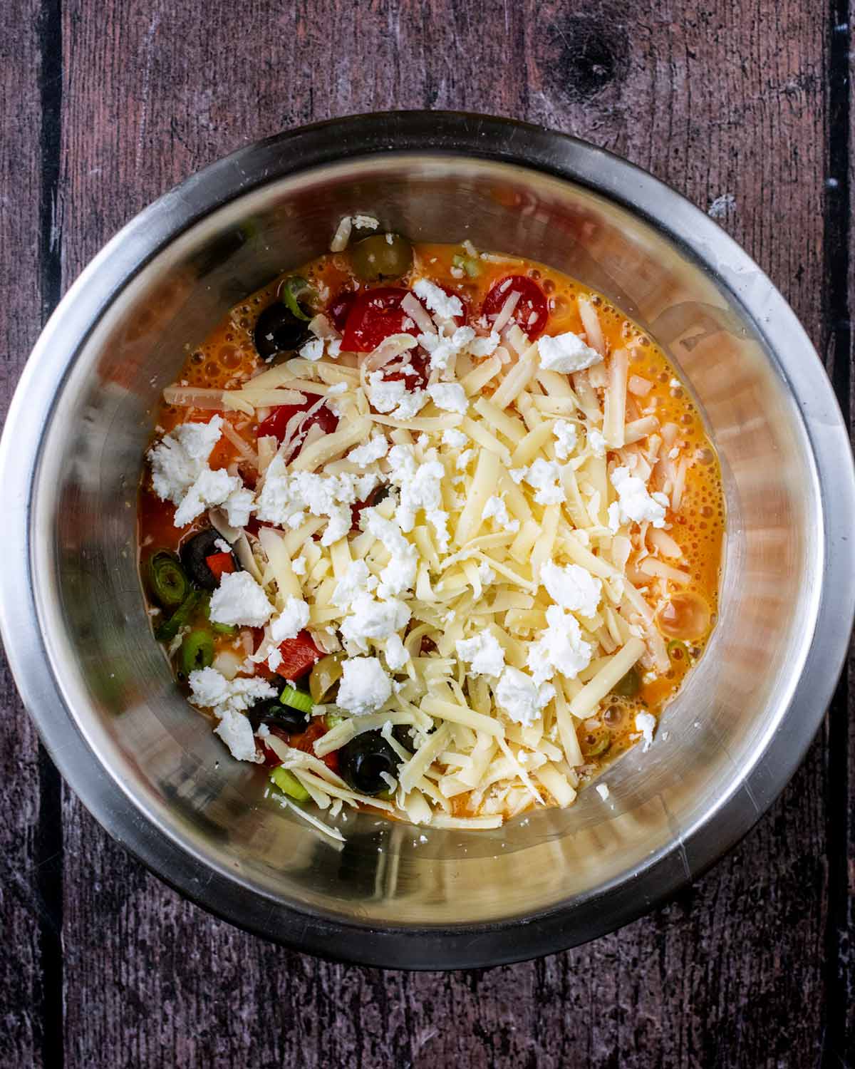 A mixing bowl containing whisked eggs, tomatoes, peppers, onions and cheese.