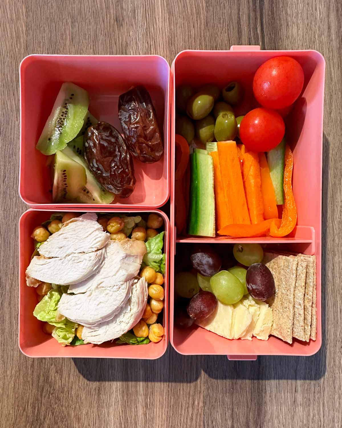 A three compartment pink bento box filled a small chicken salad, vegetables and fruit.