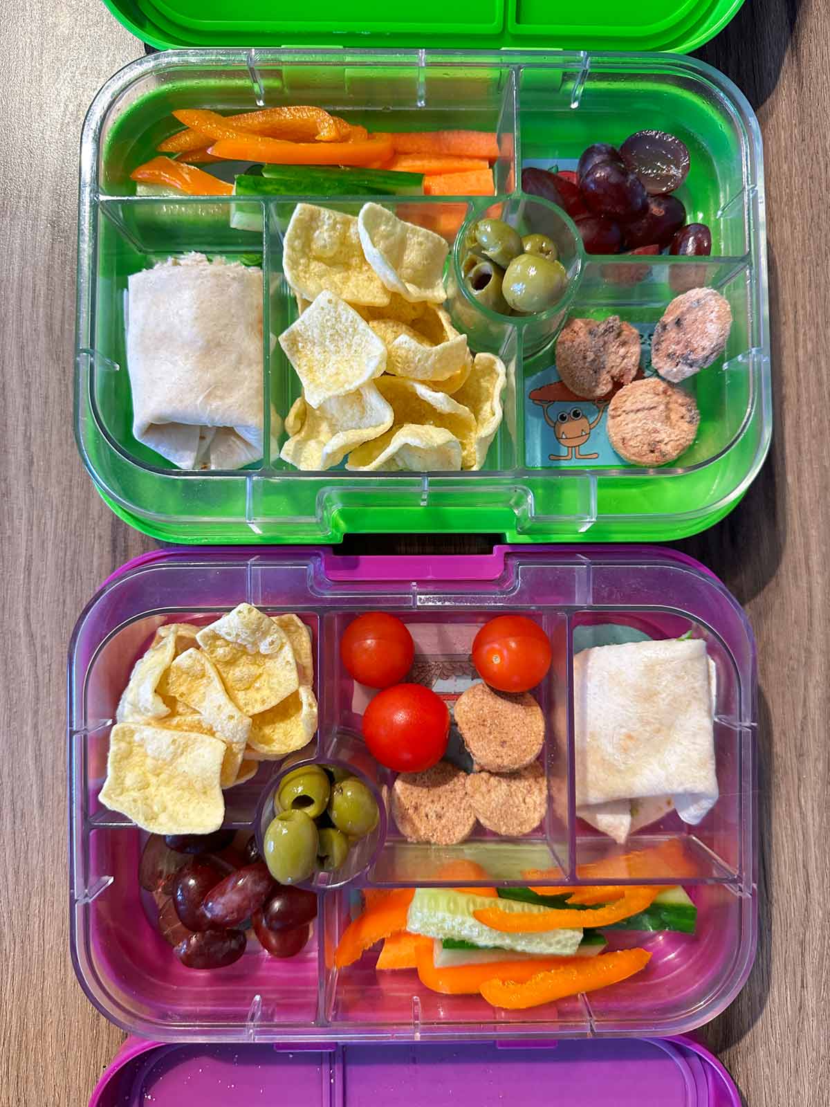 Two child's bento boxes, one purple, one green, both filled with fruit, vegetables, a wrap and mini cookies.