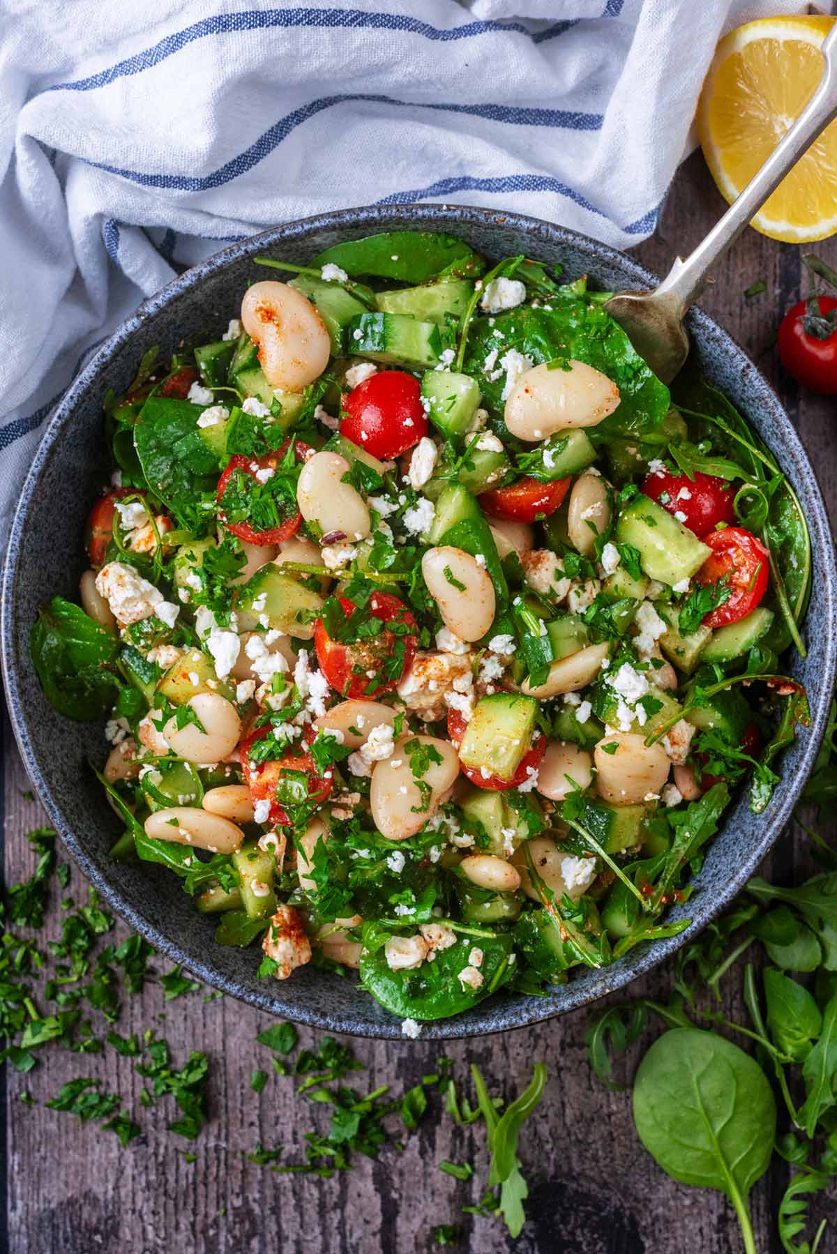 A large bowl of bean salad next to salad leaves and a lemon half.