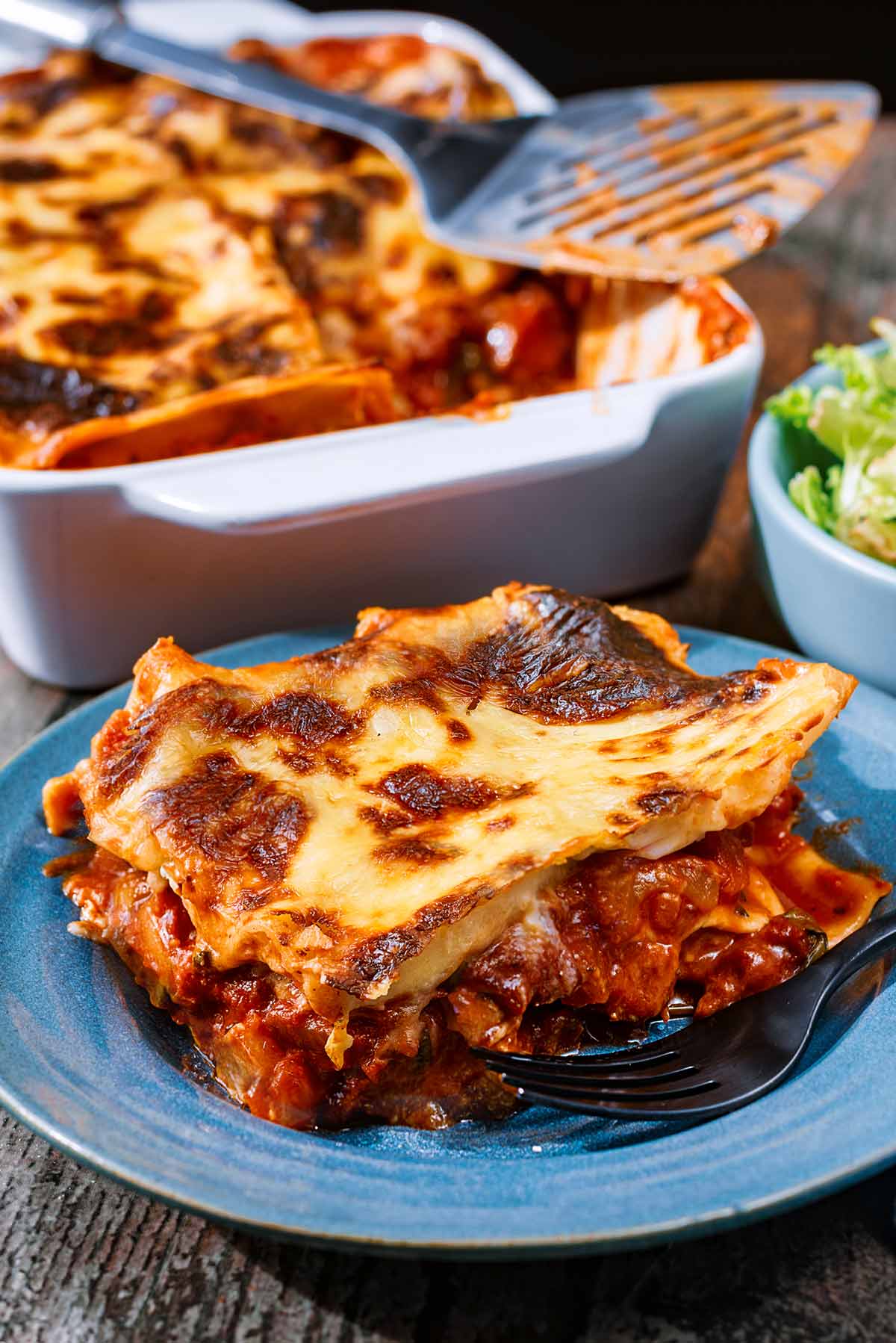 A portion of lasagna on a plate in front of a baking dish with remaining lasagna in it.