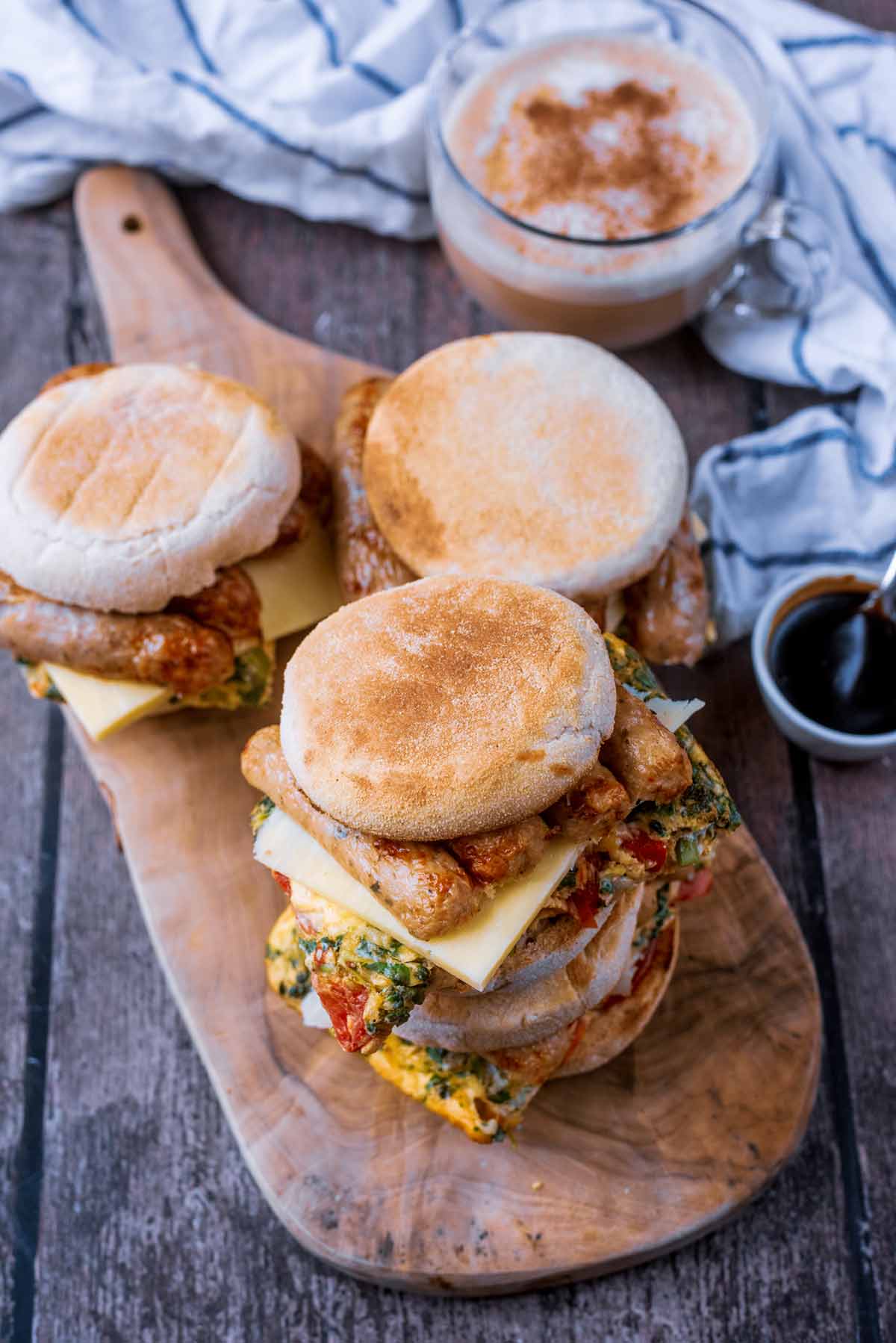 Four breakfast sandwiches on a wooden serving board next to a cup of coffee.