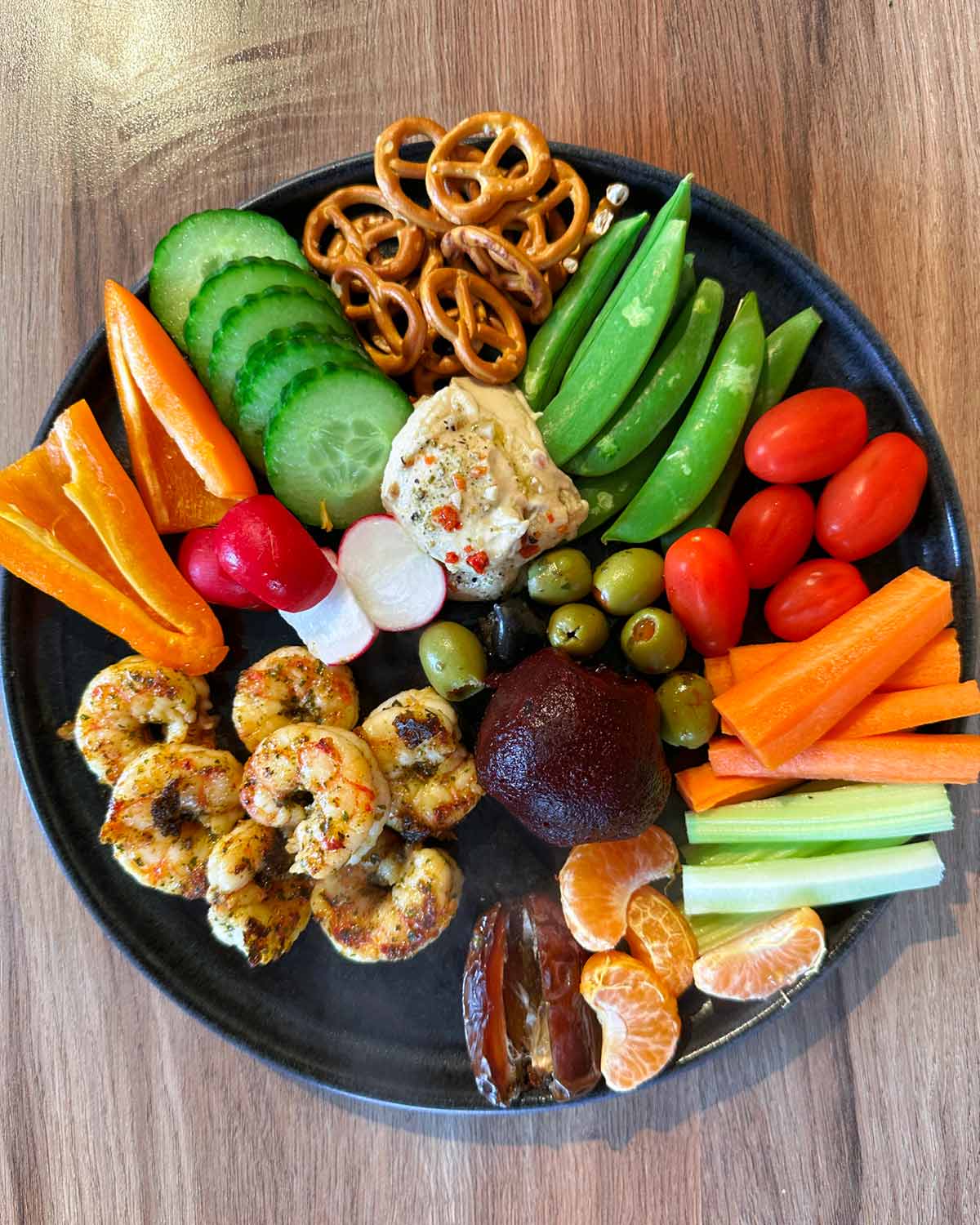 A plate of cooked prawns, carrot, cucumber, peas, tomatoes, peppers, pretzels, humus and fruit.