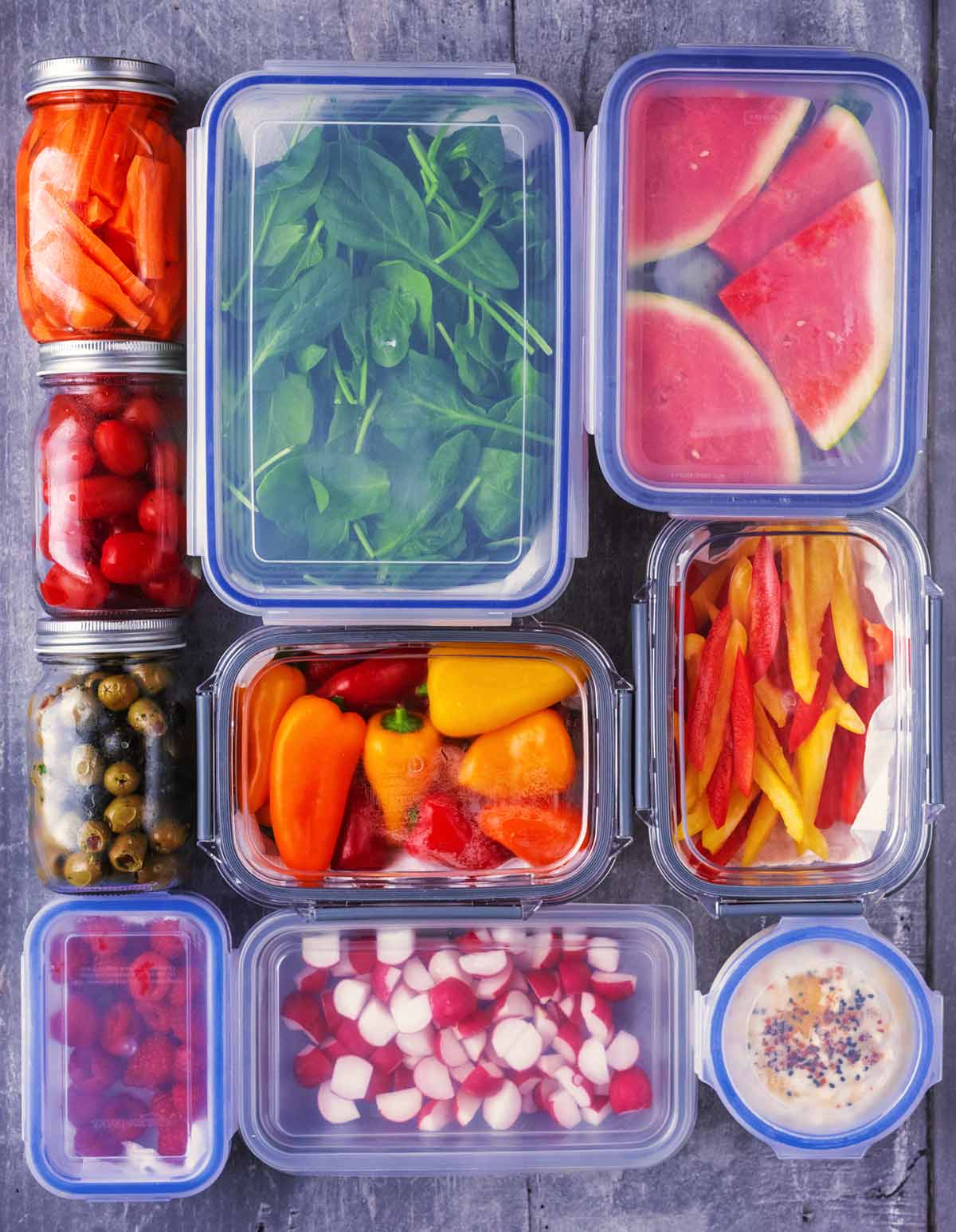 Glass containers with carrots, tomatoes, olives, berries, spinach, melon, peppers, radish and hummus.