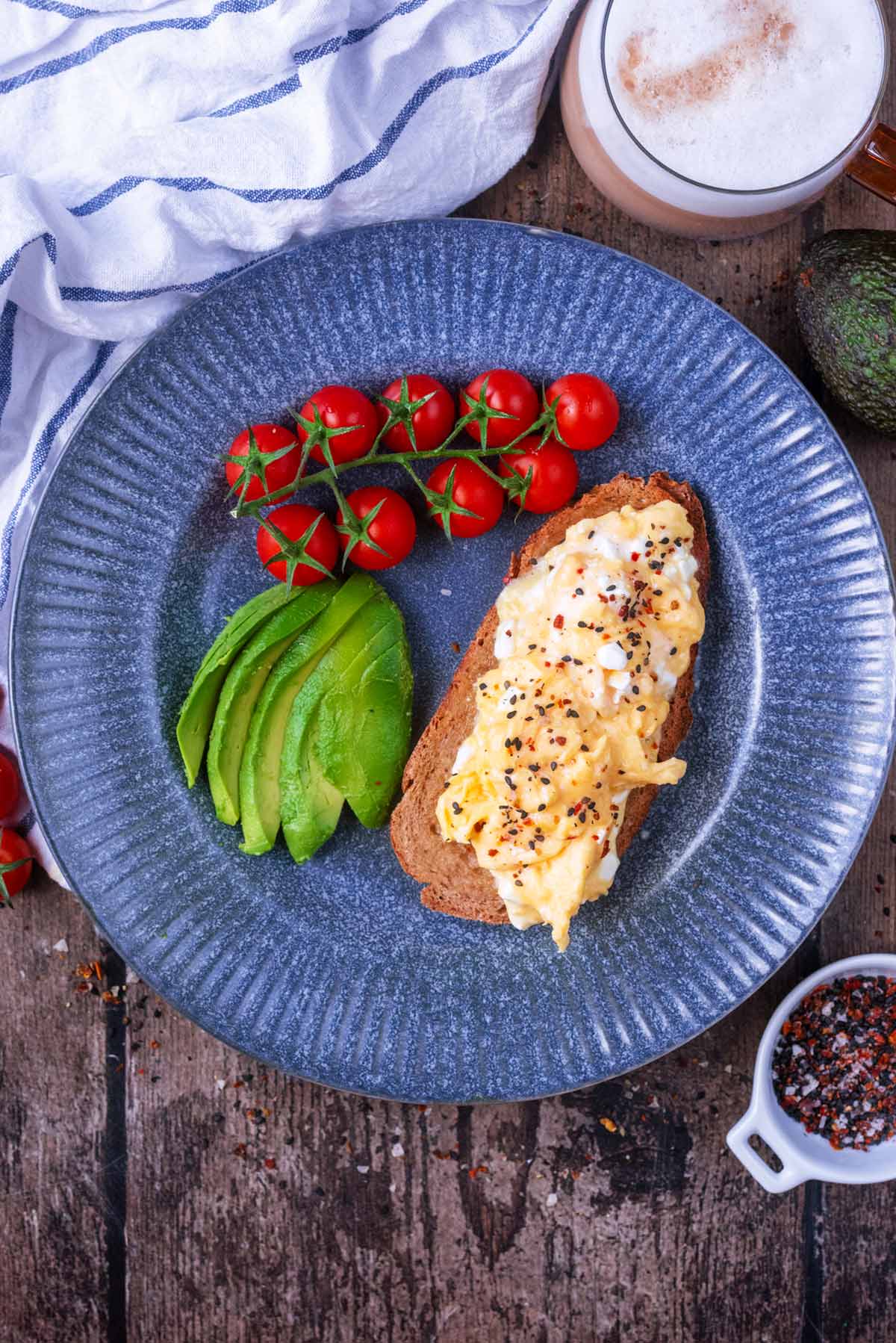 A plate of scrambled eggs on toast with cherry tomatoes and sliced avocado.